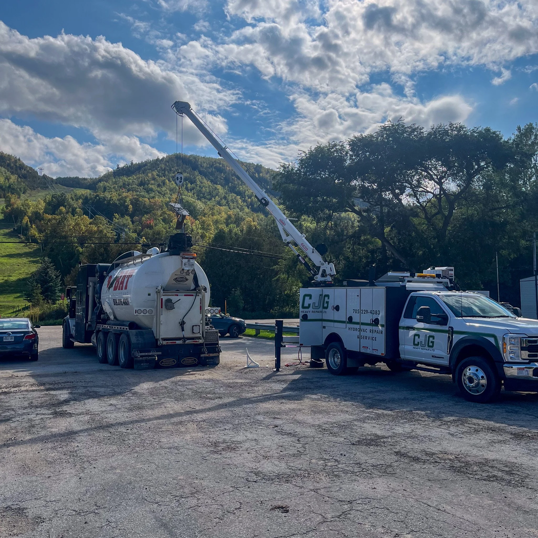 A utility truck with a crane lifting a large metal cable, parked in a gravel lot with trees and hills in the background under a partly cloudy sky.