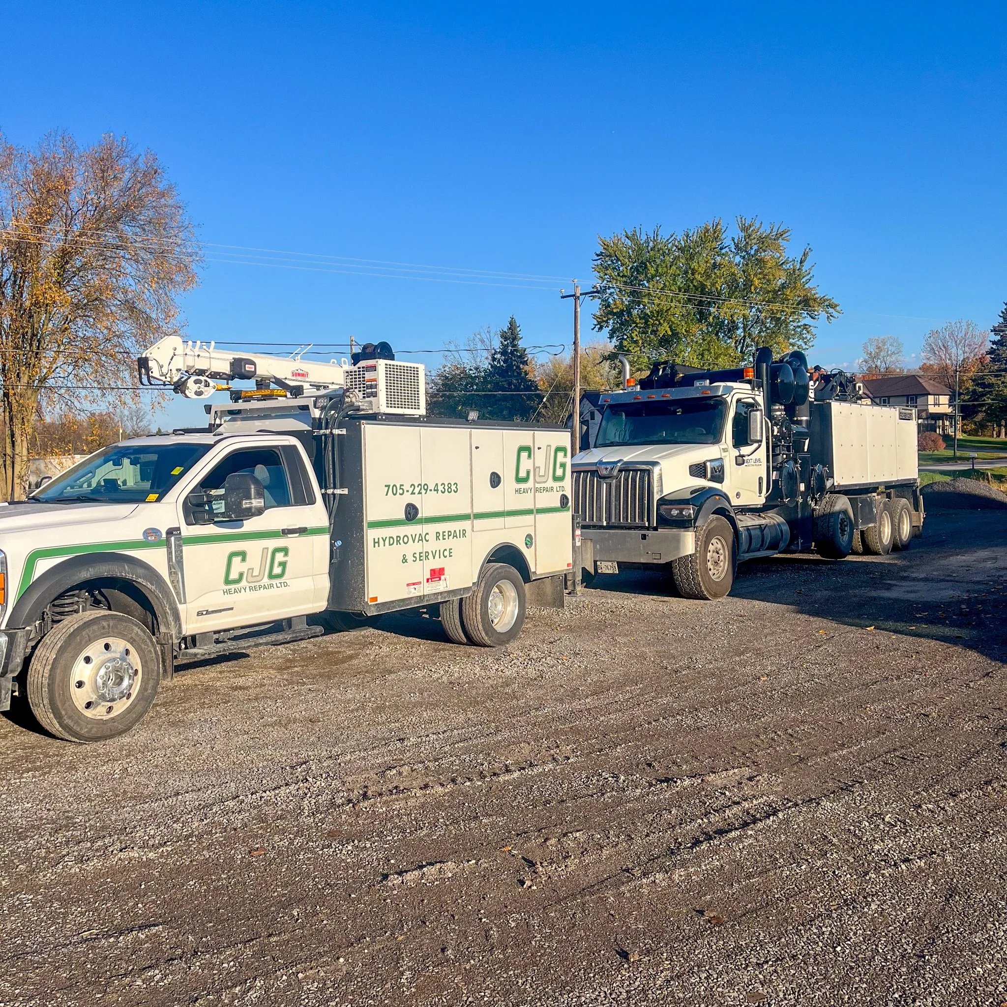 Two service trucks, one white with green accents labeled C J G Heavy Repair Ltd., and a larger black and white truck, parked on a gravel surface under a clear blue sky with trees and houses in the background.