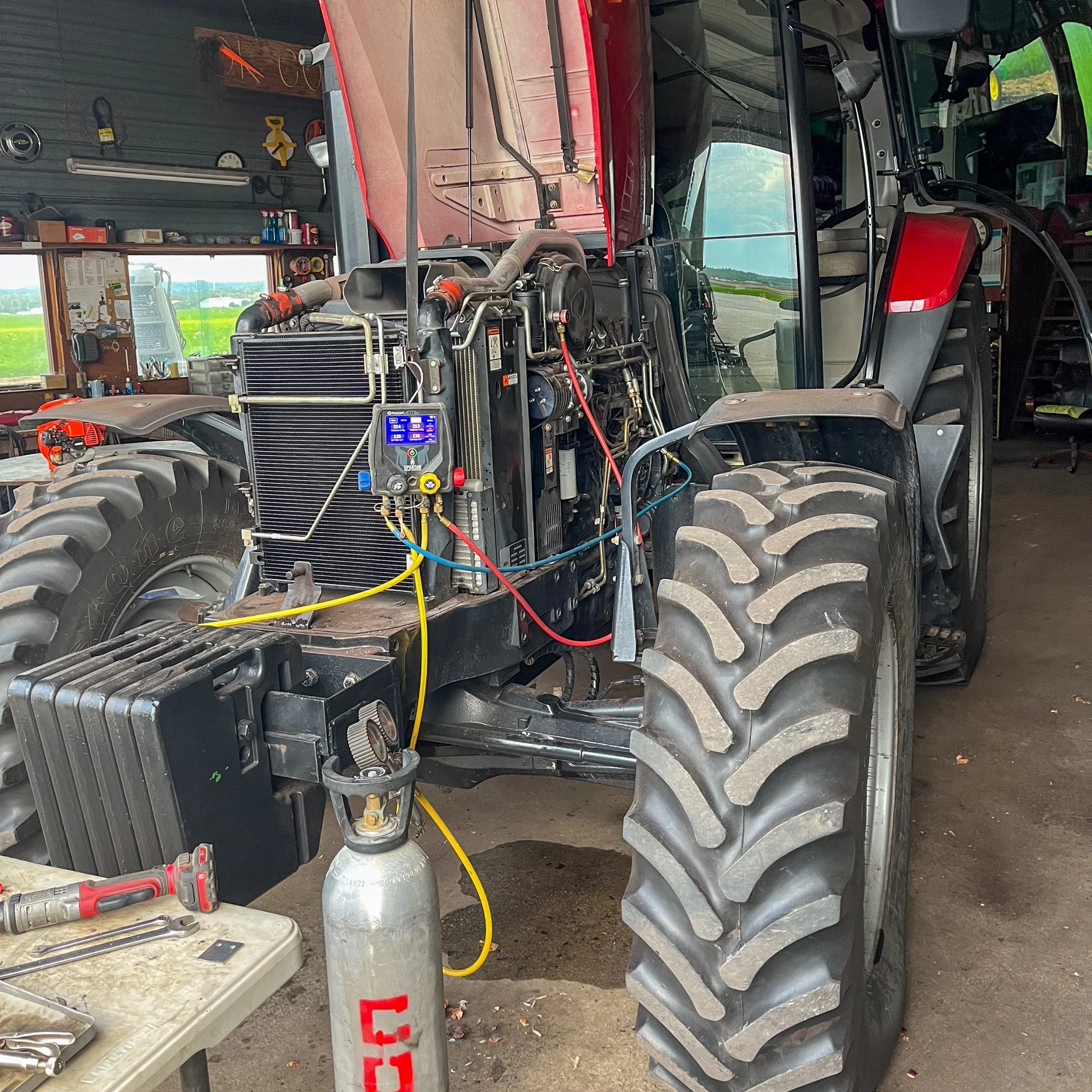 A large red tractor inside a workshop with its front panel removed, exposing the engine and mechanical components. Tools are on a nearby workbench, and the background shows a window with a view of green fields.