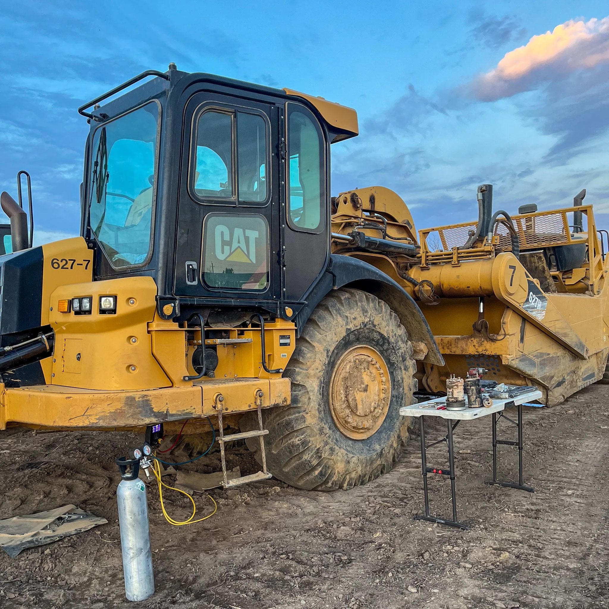 A large yellow and black CAT construction vehicle parked on dirt, with a small work table nearby holding tools and equipment. The sky above has some clouds.