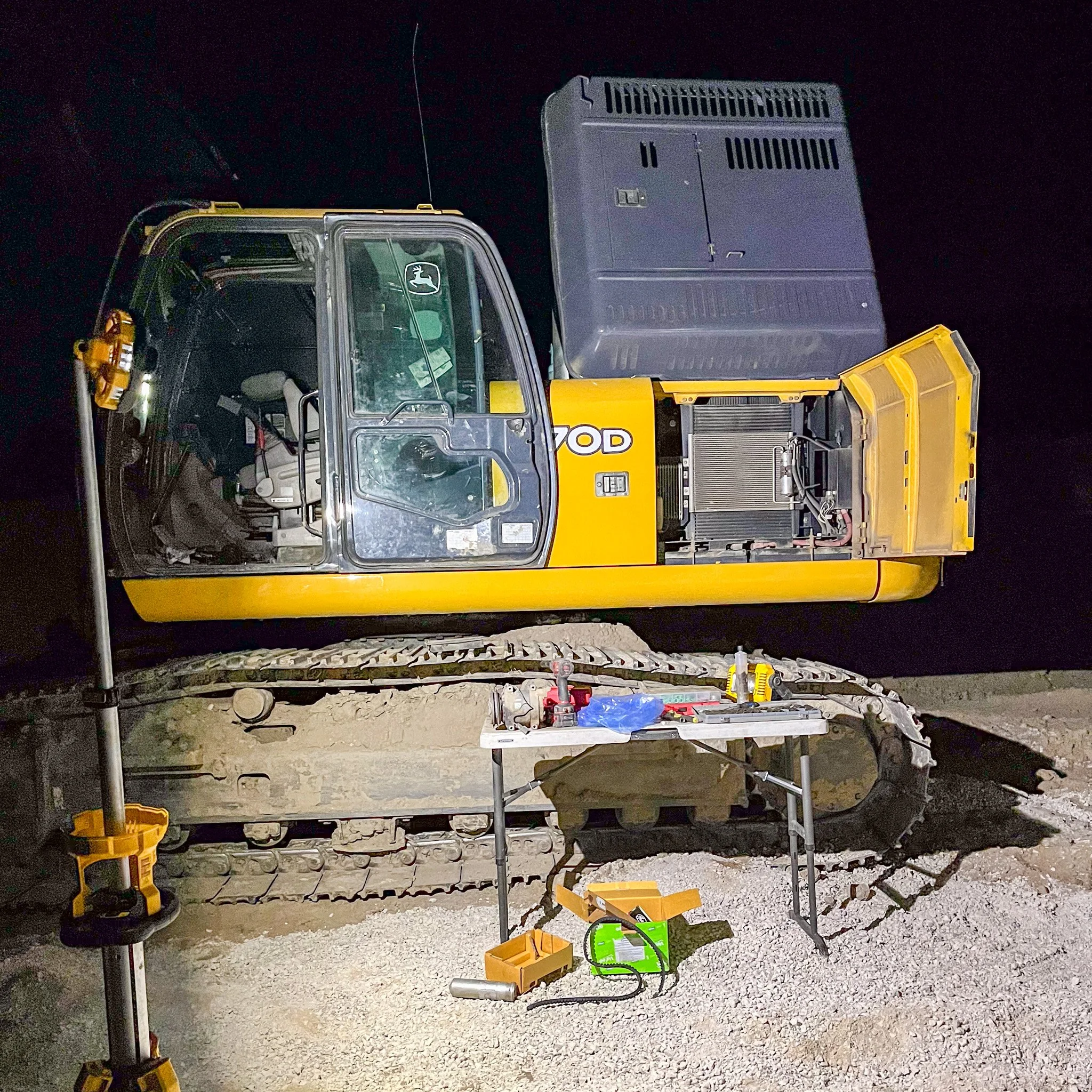 Construction site at night with a yellow John Deere excavator on dirt, surrounded by tools and equipment, illuminated by work lights.