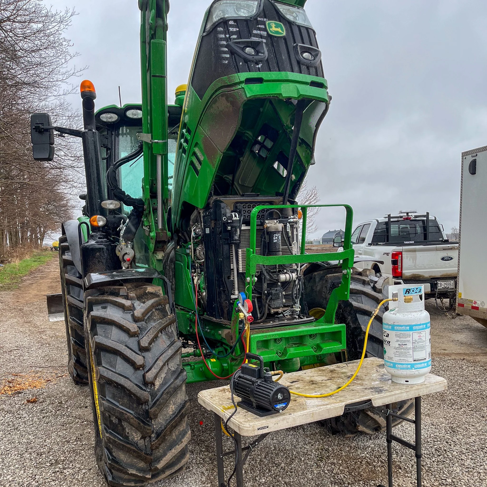 A green John Deere tractor with its engine compartment open, placed on a wooden table outdoors. There are tools and a large tank connected to the engine, with a garage or workshop trailer and pickup truck in the background under an overcast sky.