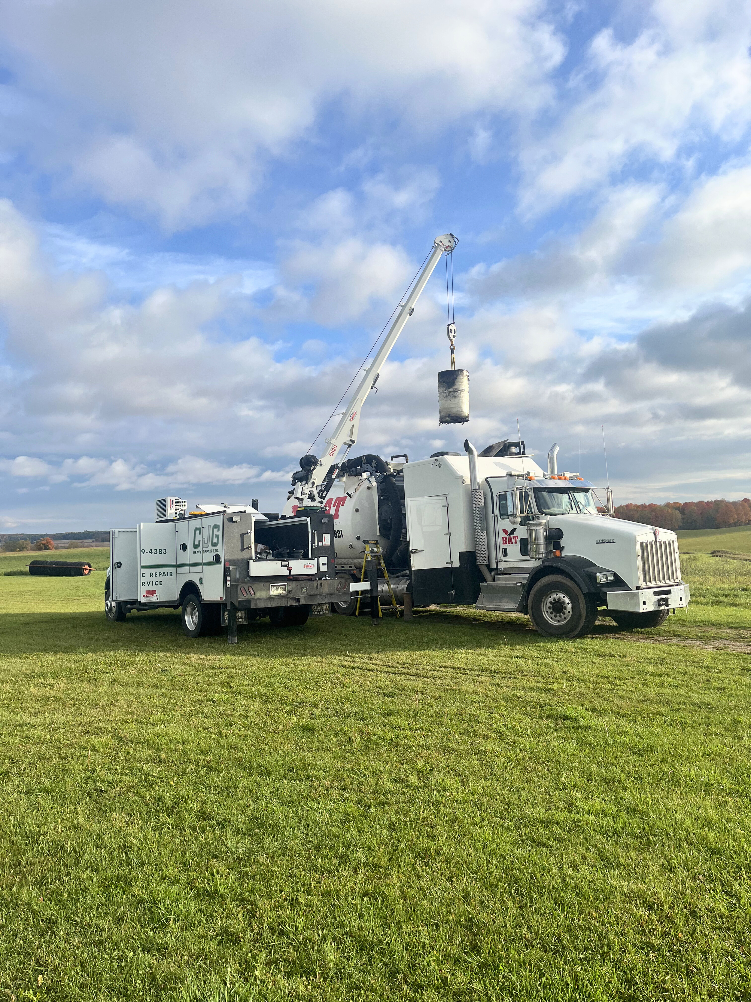 A large white tow truck with a crane is lifting a black and white barrel in a grassy field under a blue sky with clouds.