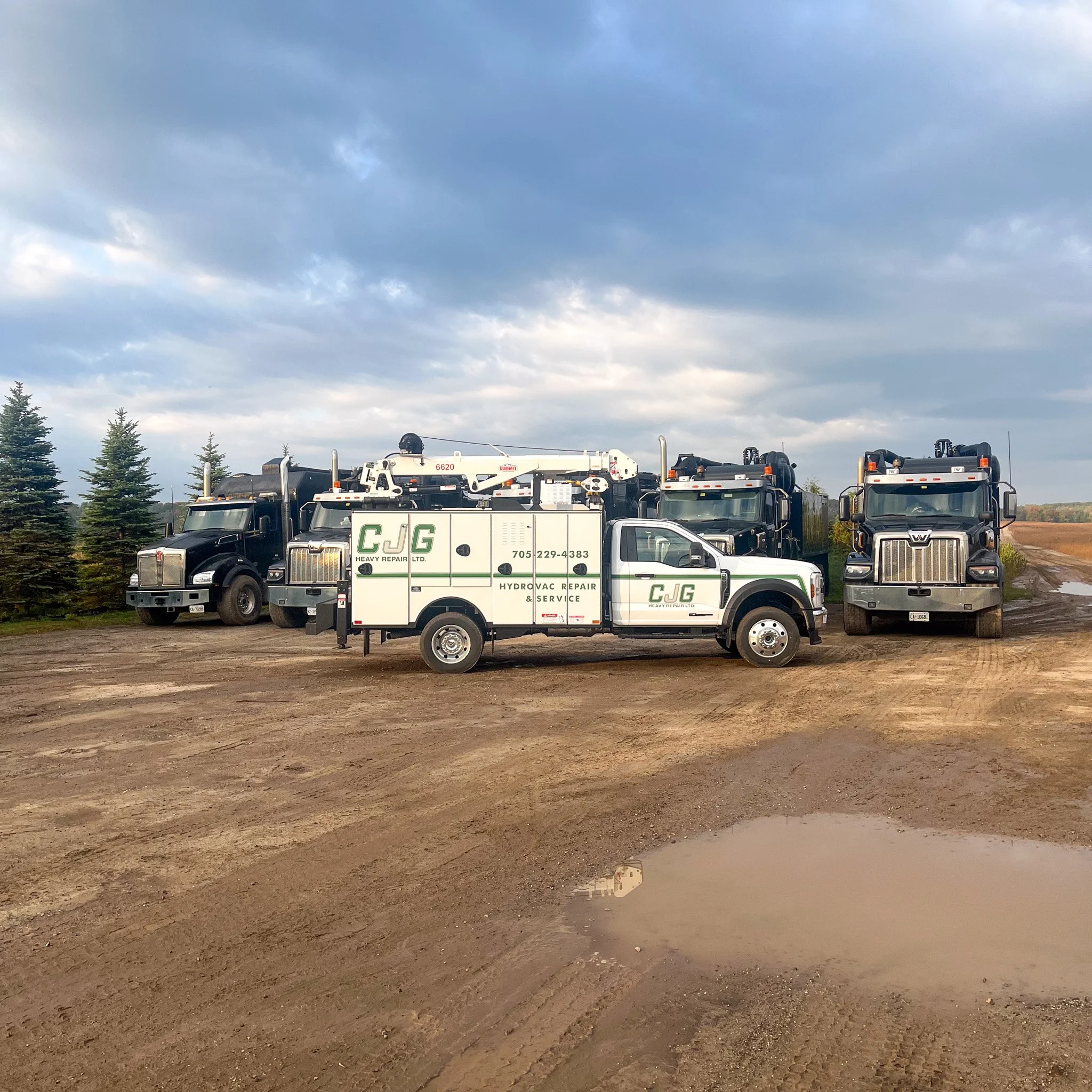 A fleet of heavy-duty repair trucks parked on a dirt lot, with a small puddle in the foreground and a row of evergreen trees in the background under a partly cloudy sky.