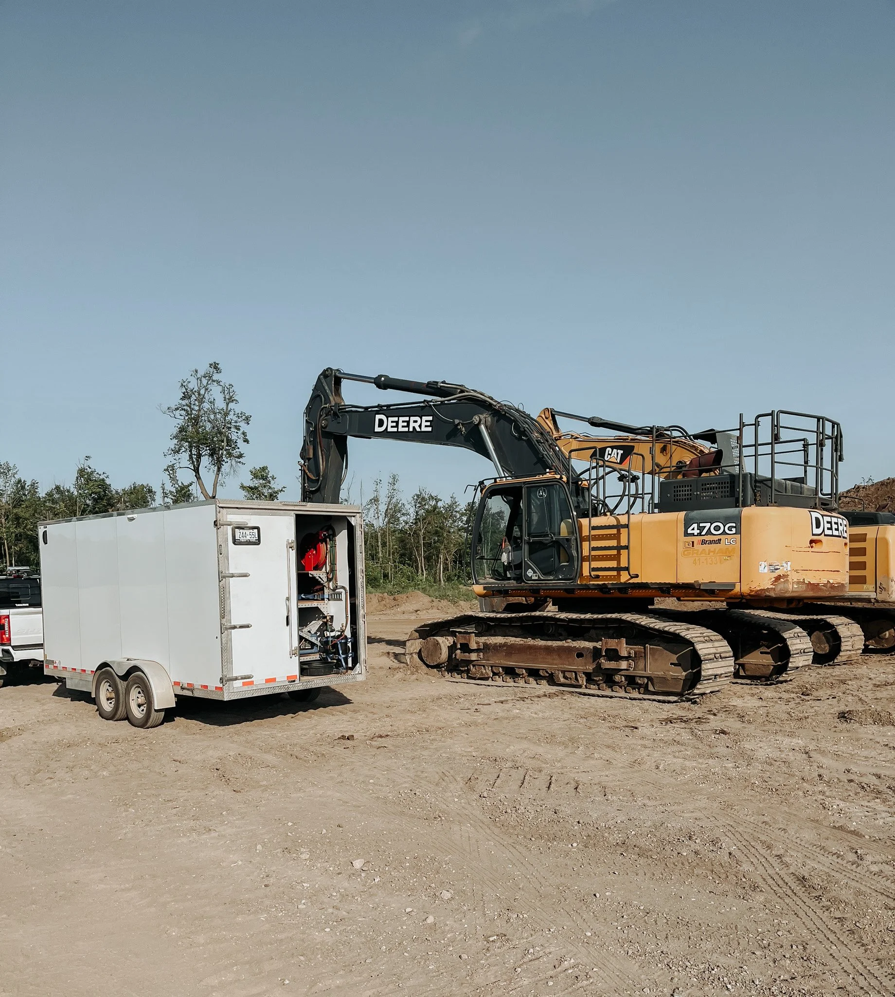 A construction site with a large yellow Deere excavator next to a white utility trailer on dirt terrain, with some trees and clear sky in the background.