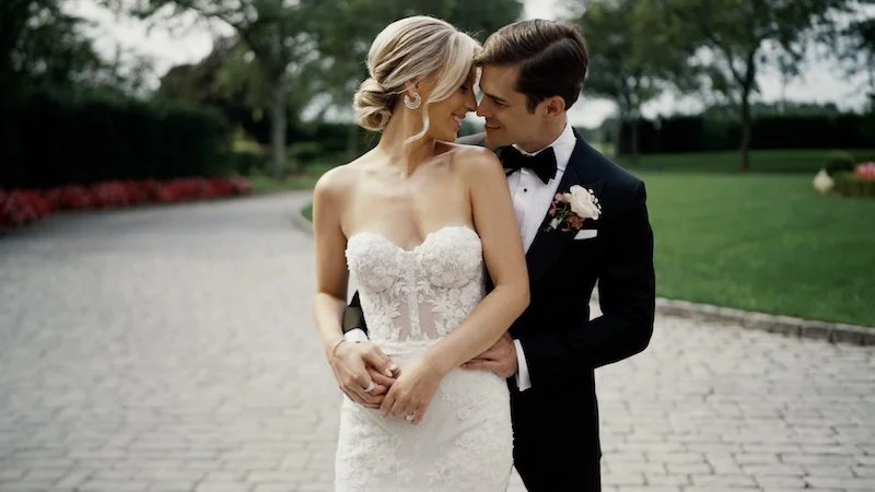 A bride and groom smiling and embracing each other outdoors on their wedding day, with a garden and trees in the background.