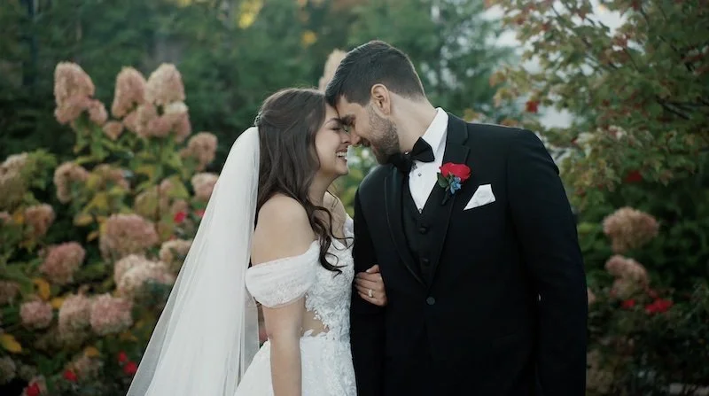 Bride and groom touching foreheads and smiling outdoors, with pink flowers and greenery in the background.