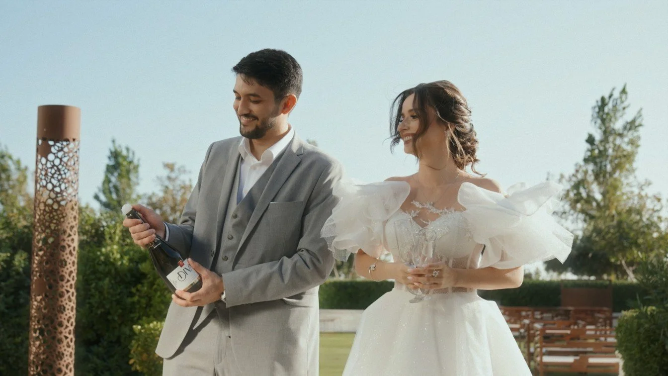 A man in a gray suit opening a bottle of champagne while a woman in a white wedding dress with large ruffled sleeves holds a champagne glass, outdoors on a sunny day.