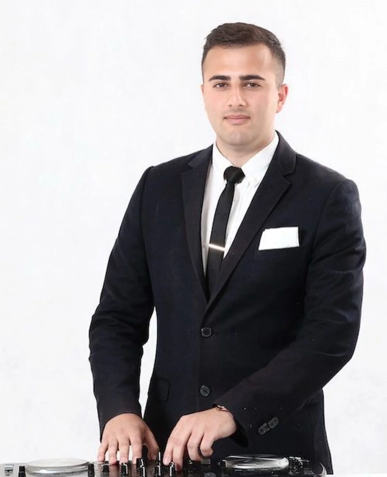 A young man in a black suit and white shirt wearing a black tie, standing with hands on DJ equipment against a plain white background.