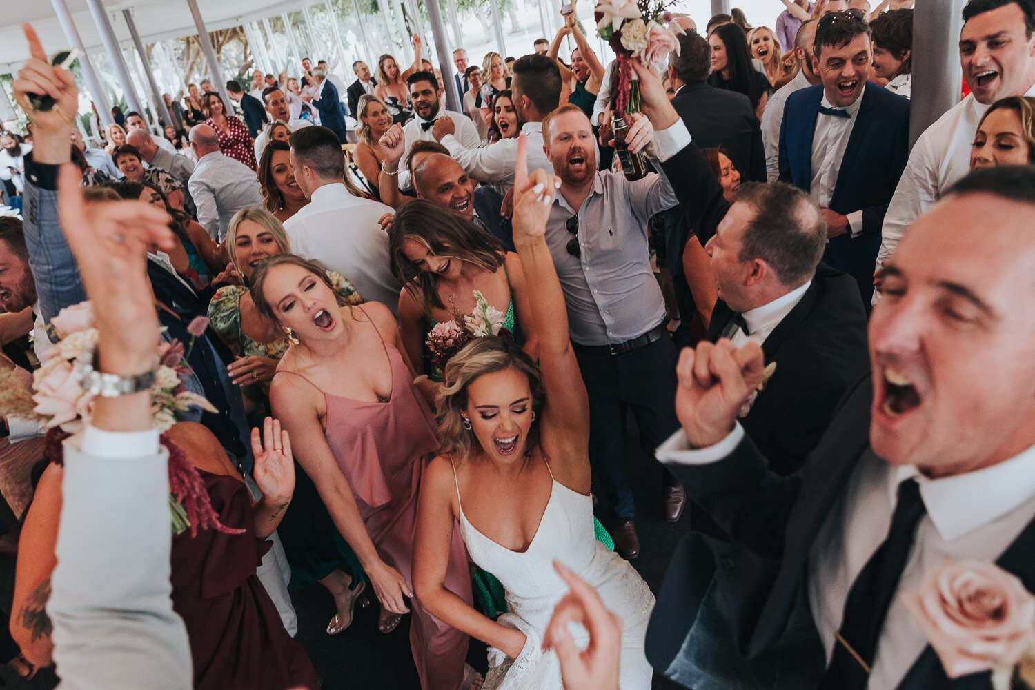 People dancing and celebrating at a wedding reception, smiling, singing, and raising their hands in a lively atmosphere.