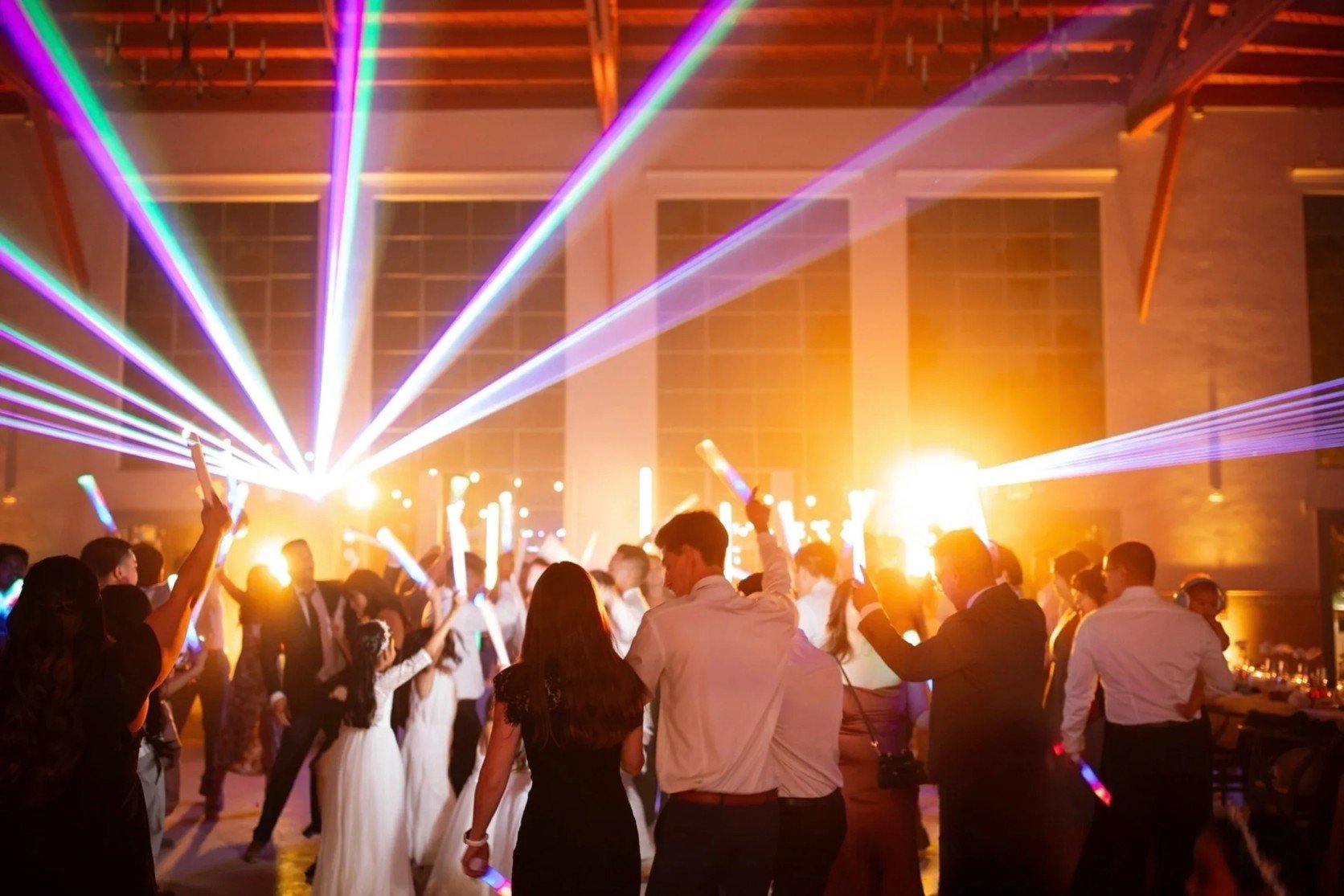 People dancing at a party with colorful laser lights and glow sticks in a large indoor venue.