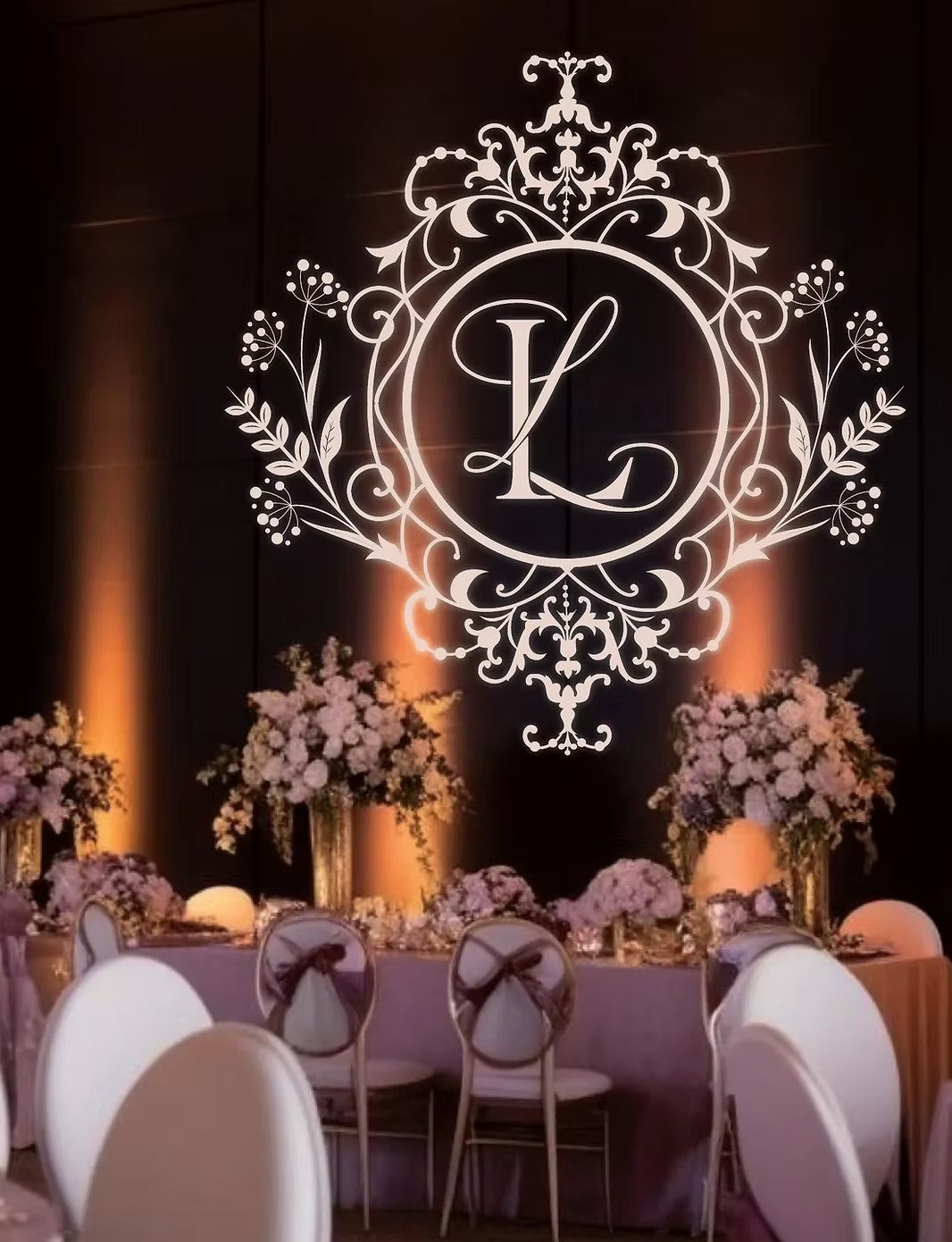 Elegant event decor with a black wall featuring a white monogram logo with the initials 'L' and 'K' surrounded by ornate floral designs and illuminated by soft lighting. A table with white chairs, decorated with pink and lavender floral arrangements and elegant centerpieces, is set in front of the wall.