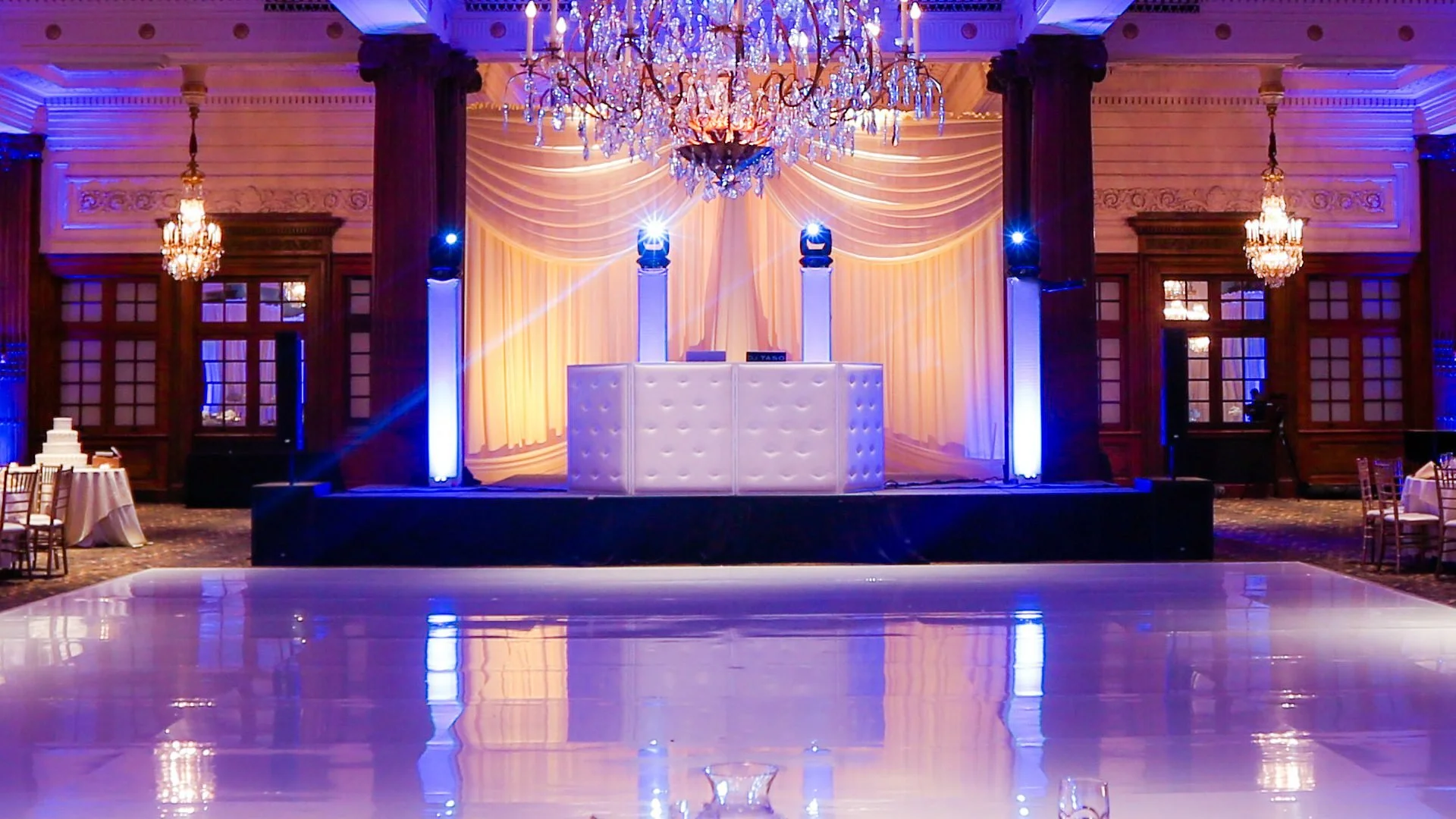 An elegant event venue with a dance floor, decorated with chandeliers and warm lighting, featuring a DJ booth on a stage with a curtain backdrop.