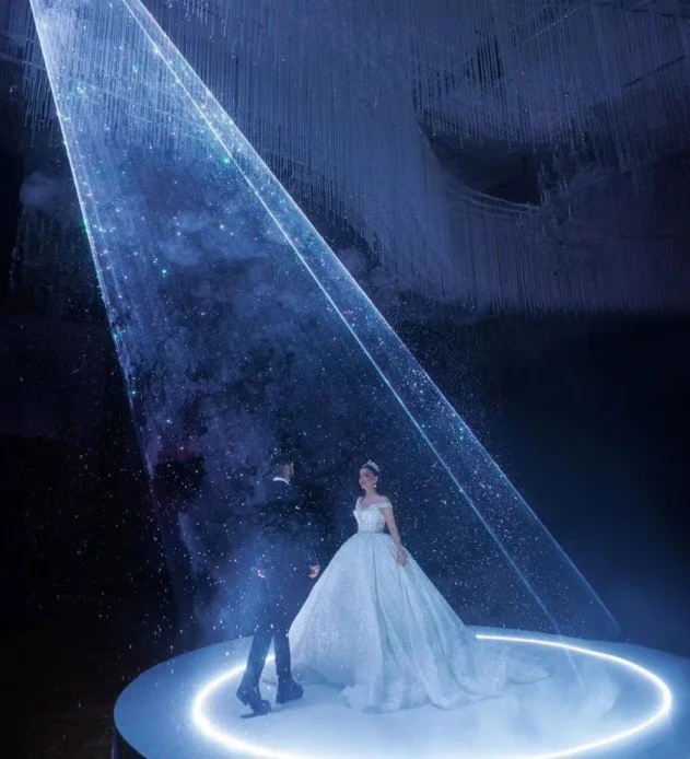 A couple dressed in formal wedding attire standing on a circular platform with futuristic, holographic light effects creating a starry, cosmic scene.