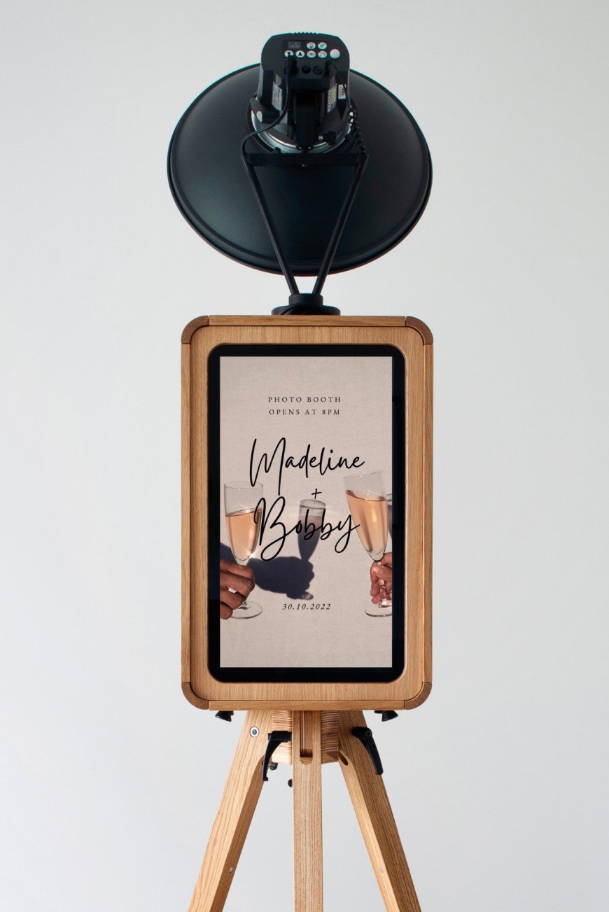 Photo booth setup with a digital screen inside a wooden frame on a tripod and a camera mounted above it. The screen displays text about a photo booth event for Madeline and Bobby on October 30, 2022.