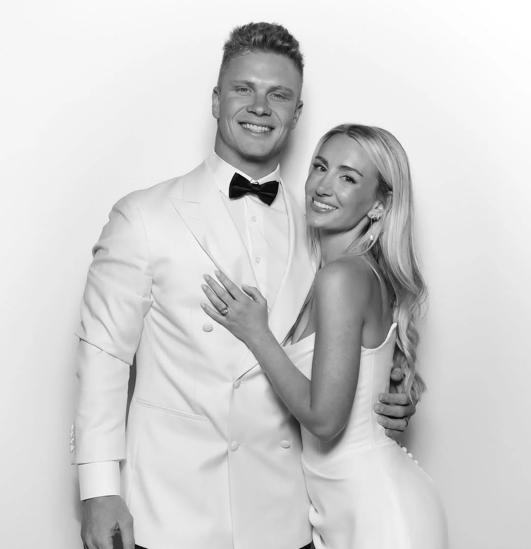 A happy couple dressed in formal attire, embracing and smiling at the camera against a plain background.