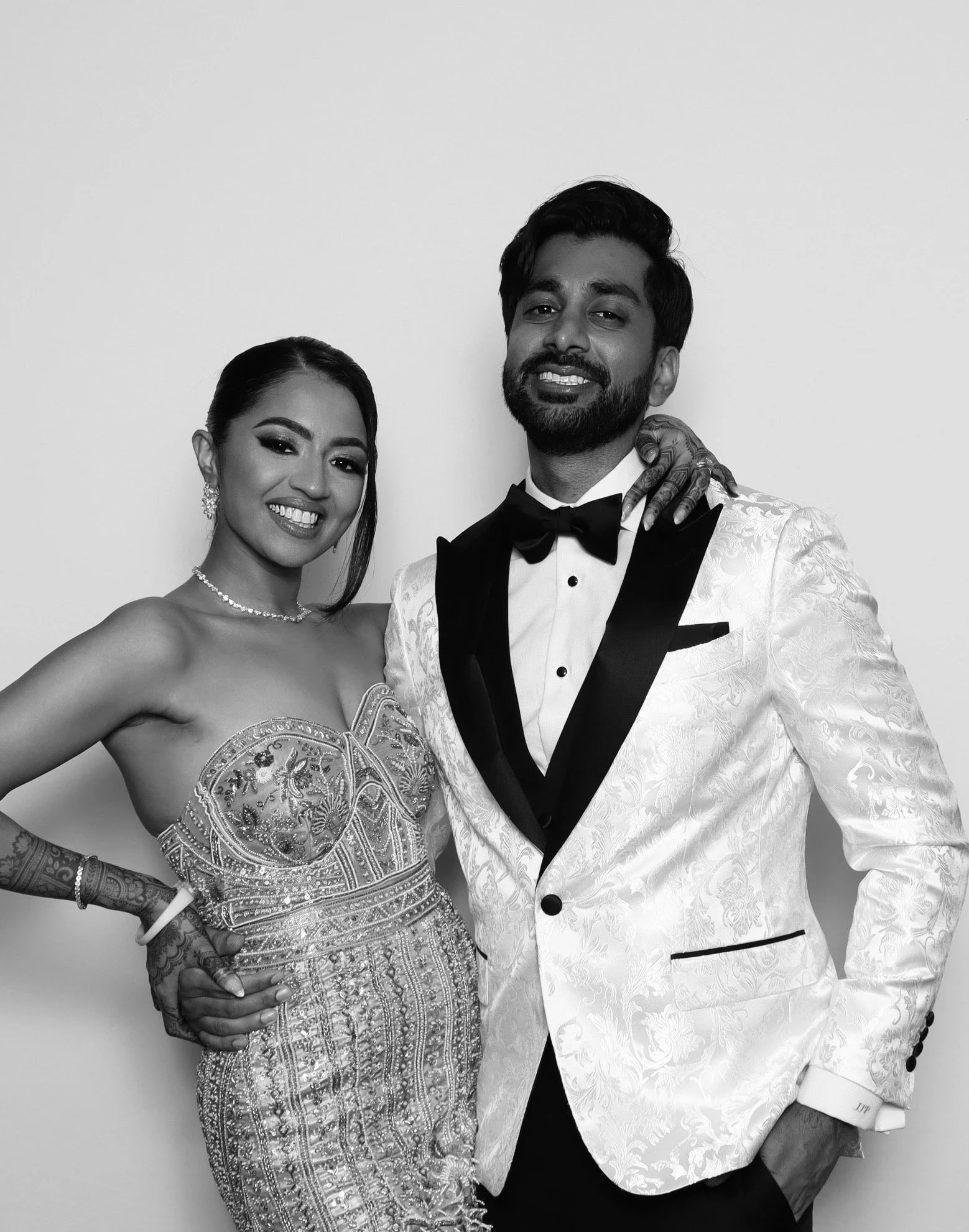 A smiling woman in a detailed, beaded gown and a man in a tuxedo with a white jacket and black bow tie, posing together against a plain background.
