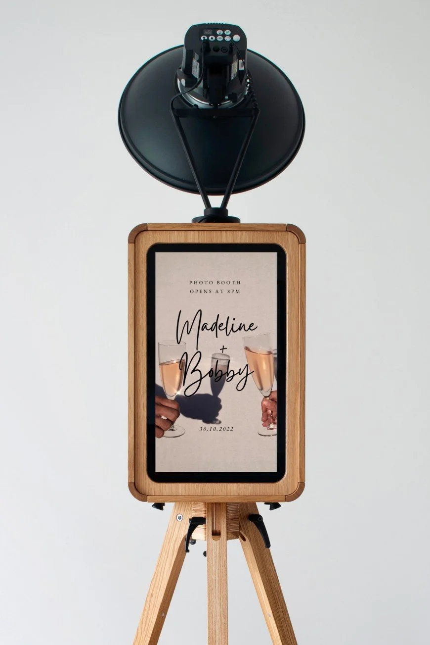 A photo booth with a wooden frame and tripod stand, displaying a sign with the names Madeline and Bobby, wedding date 30.10.2022, and people holding champagne glasses in the background.