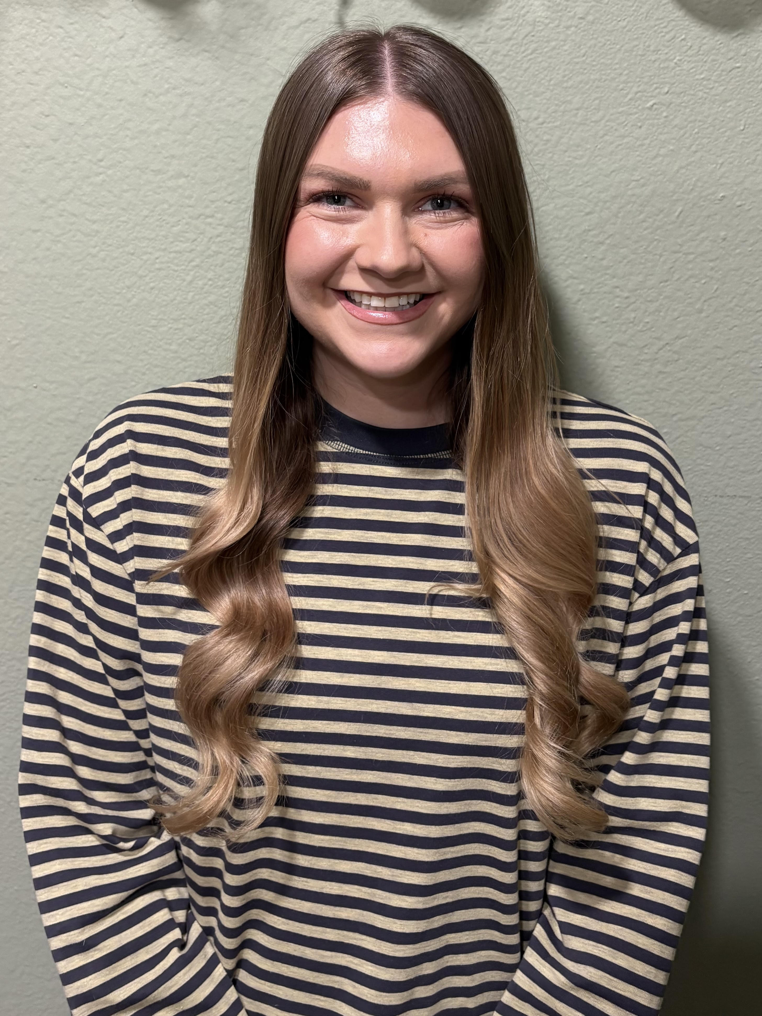 A woman with long, wavy, light brown hair and fair skin, smiling, wearing a black and beige striped shirt, standing against a plain light green wall.