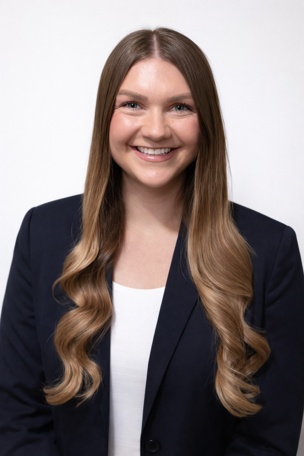 A smiling woman with long, wavy brown hair wearing a black blazer and white top against a plain white background.