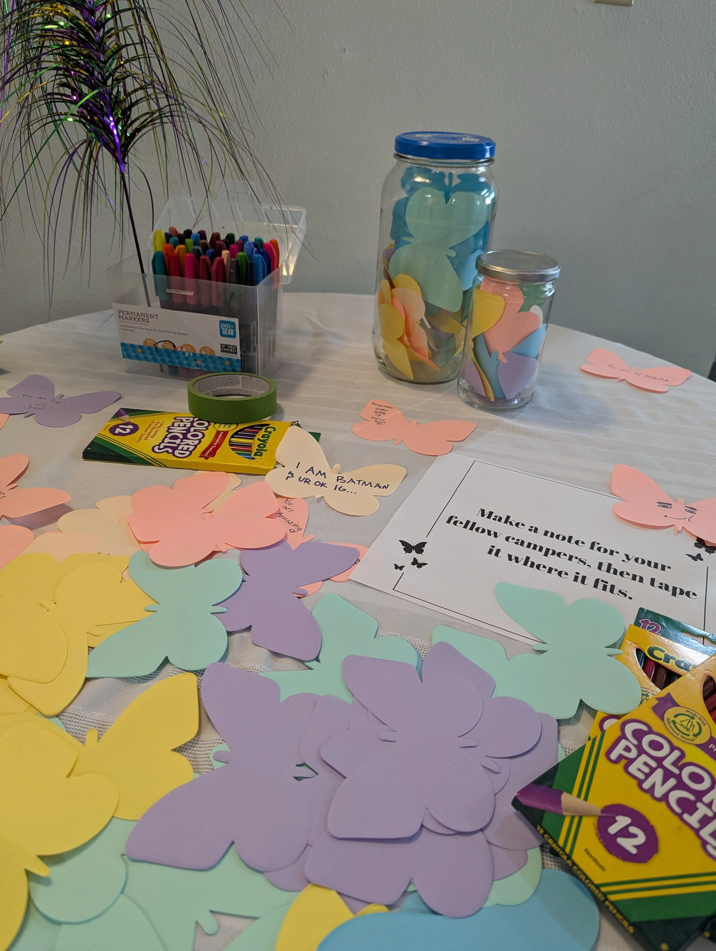 Craft table with paper butterfly cutouts in pastel colors, colored markers, tape, crayons, jars filled with butterfly-shaped paper, and a sign with instructions for making a note for campers.