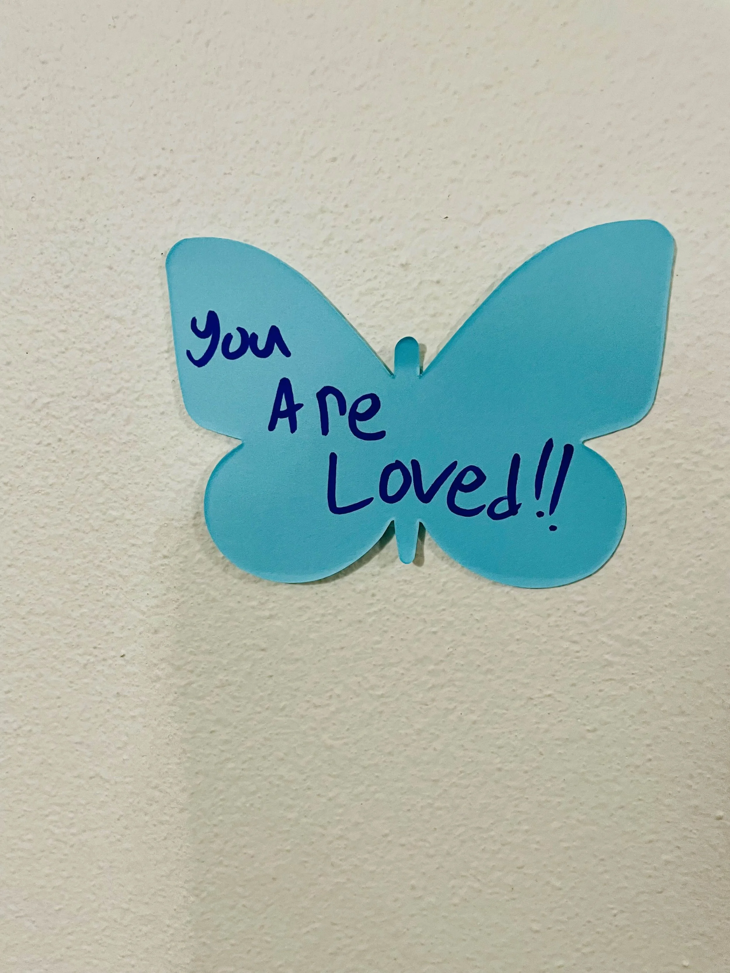 Blue paper butterfly with the handwritten message 'You Are Loved!!' on it, attached to a white wall.