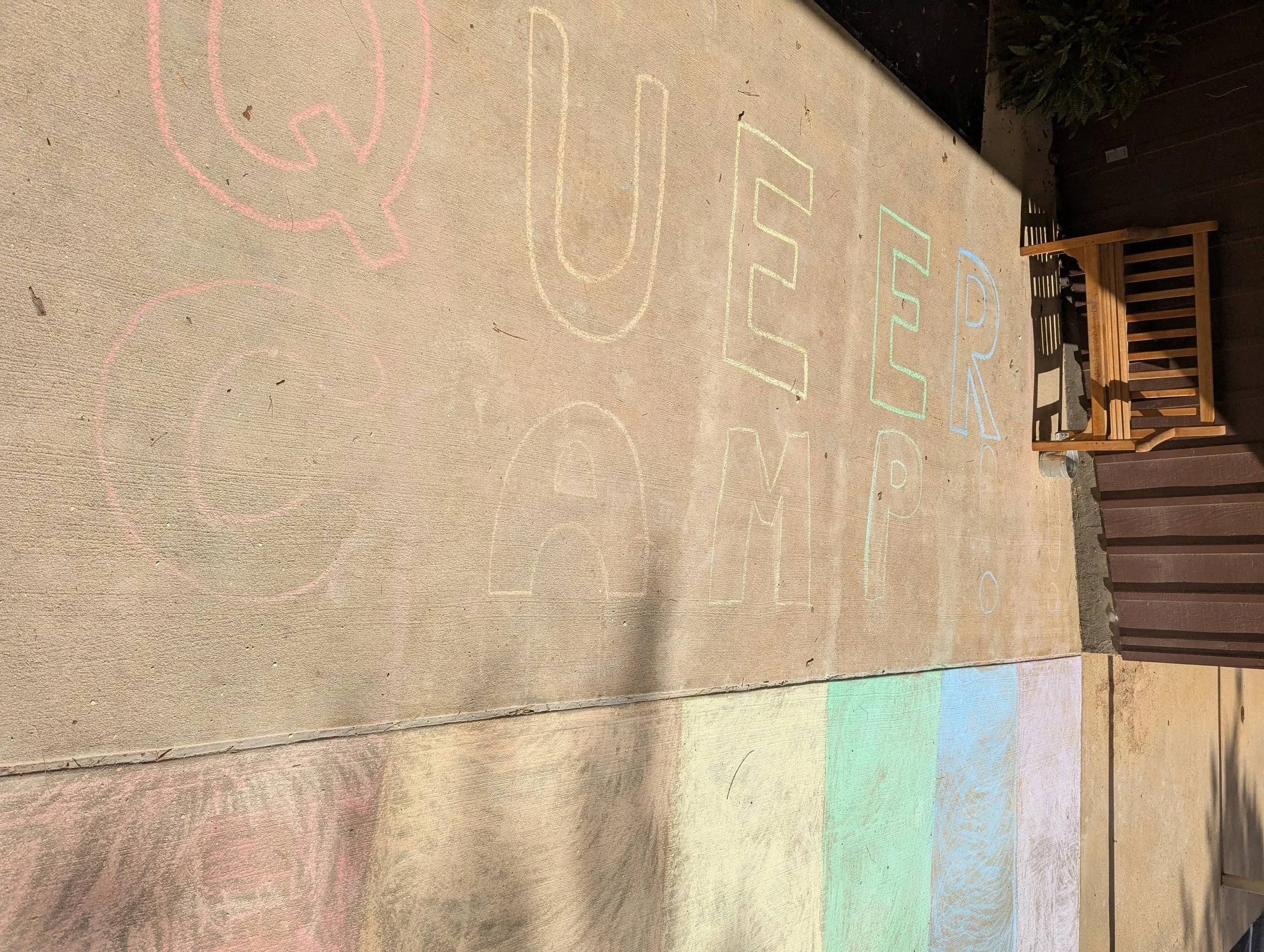 Colorful chalk message on sidewalk that reads 'QUEER CAMP' with a wooden bench nearby.
