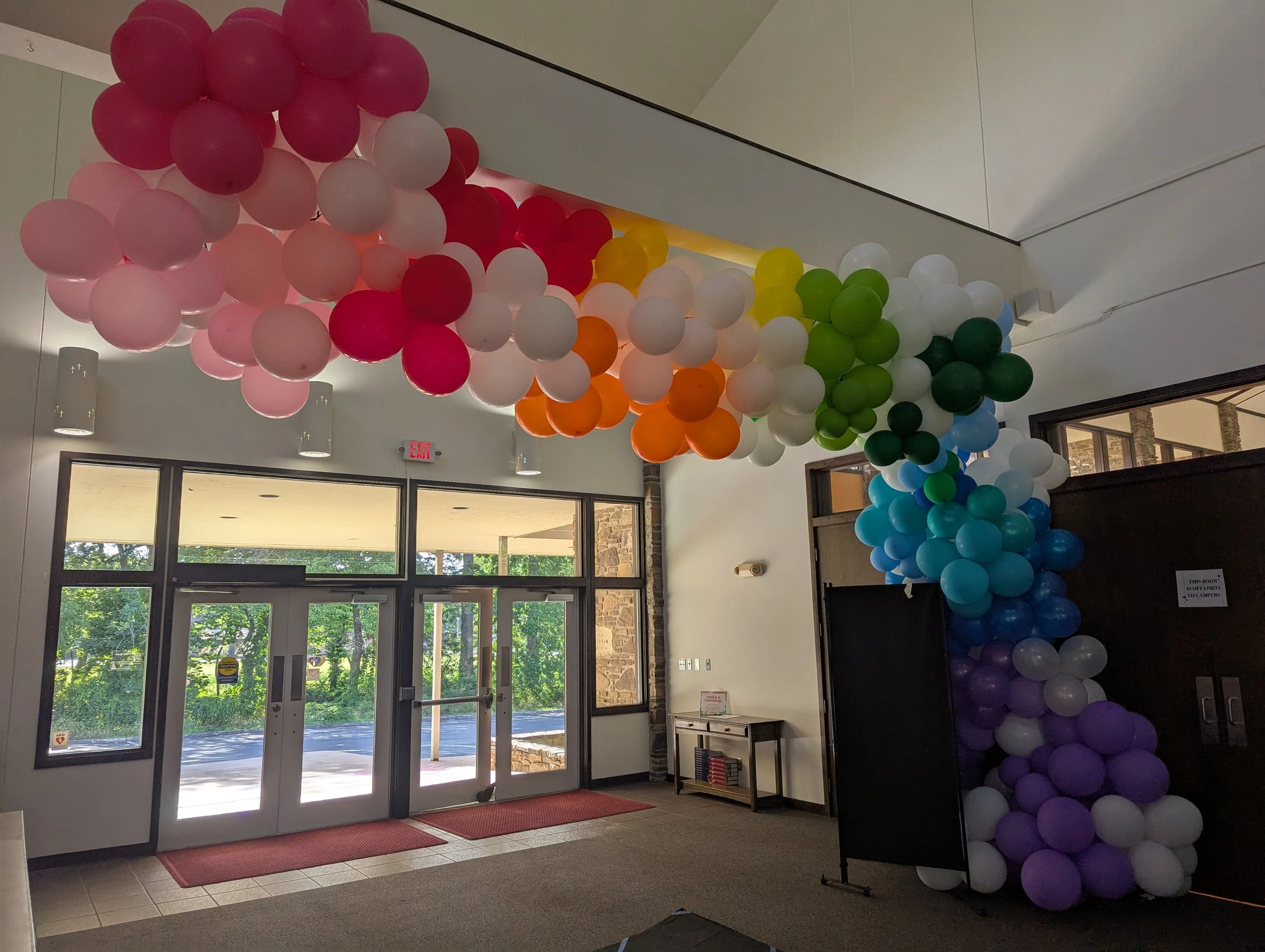 Colorful rainbow balloon arch inside a building near glass doors, with green, blue, purple, and other colored balloons arranged in an arc.