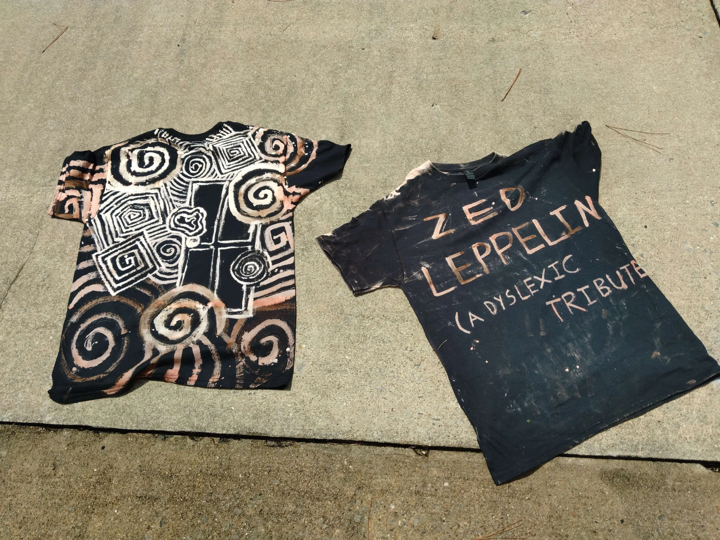 Two tie-dye T-shirts lying on concrete sidewalk, one with black and pink swirling patterns and the other with black background and pink text reading 'ZED LEPELLIN (A DISLEXIC TRIBUTE)'.
