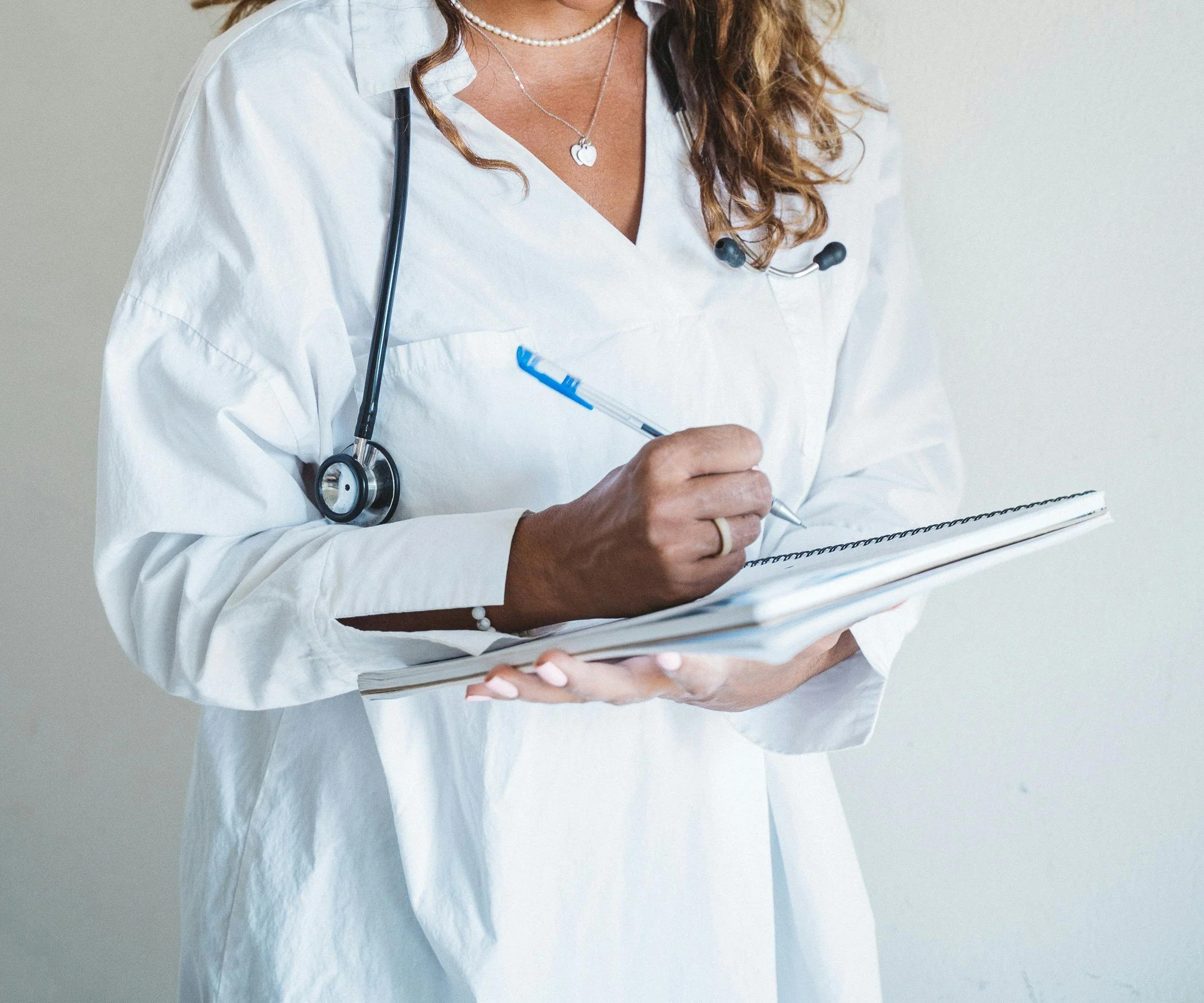 A healthcare professional in a white coat and a stethoscope around her neck is holding a clipboard and writing with a pen.