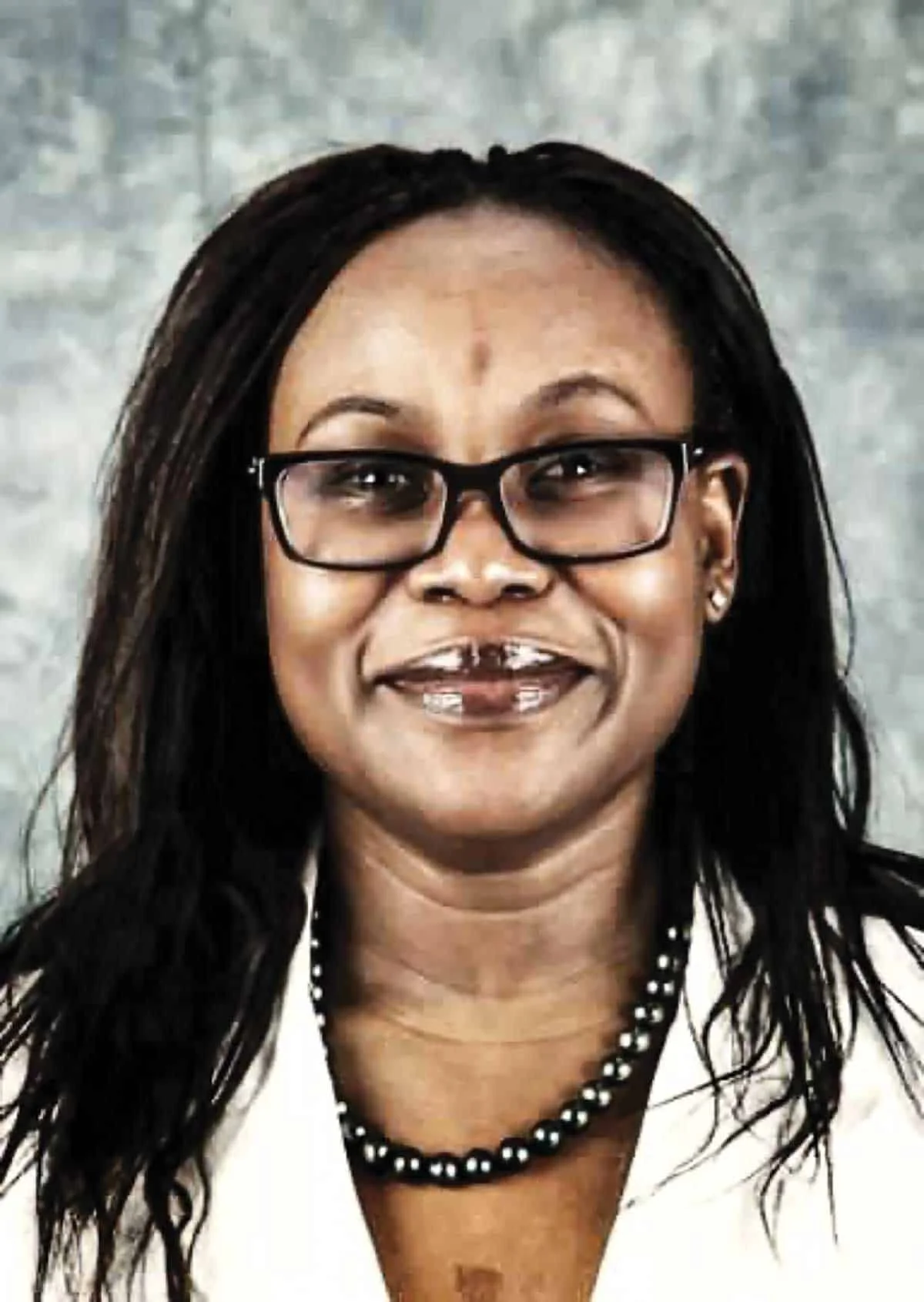 A woman with dark hair, glasses, and a beaded necklace smiling in front of a gray textured background.