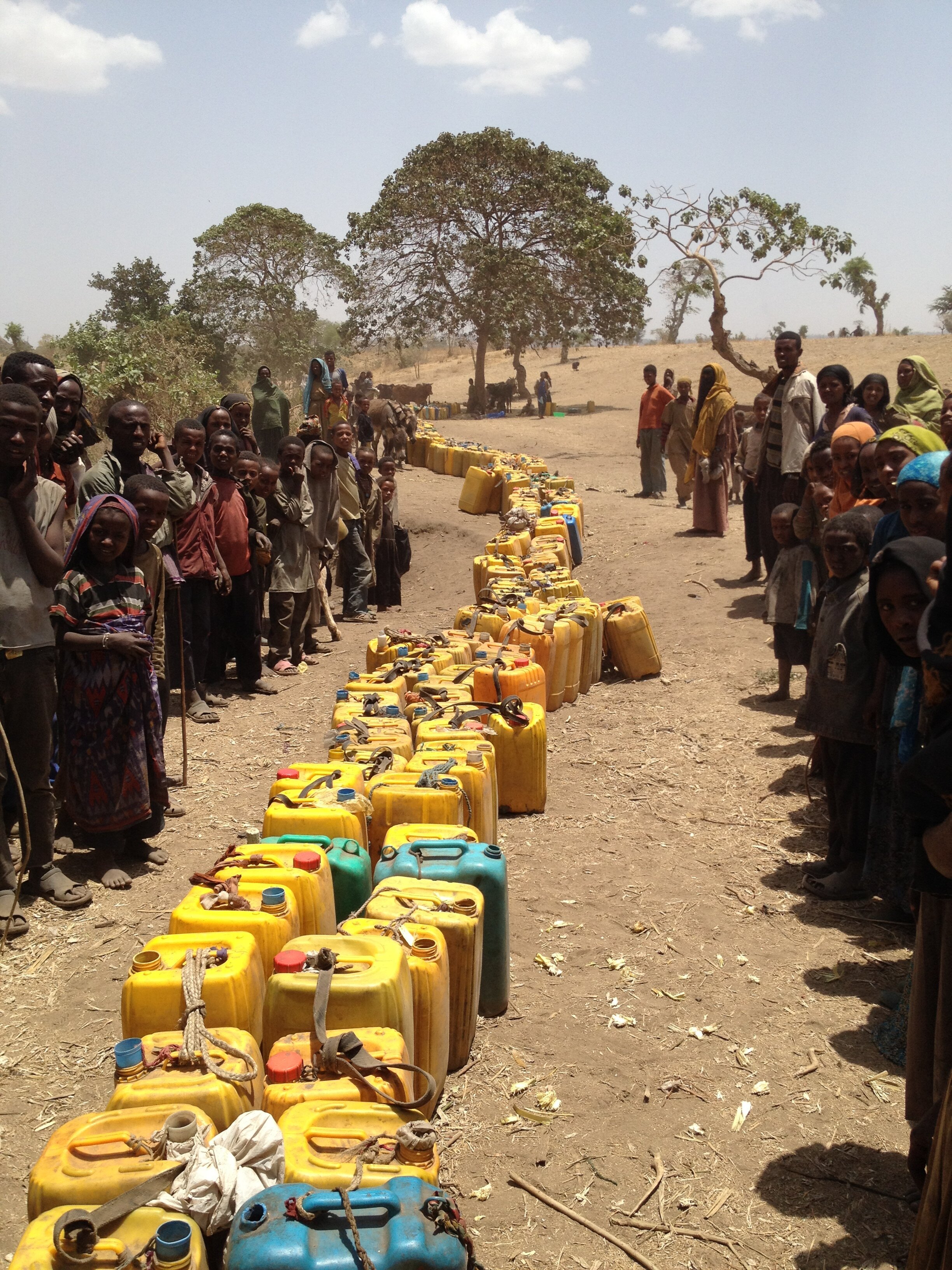 Children and adults stand in line on either side of a line of yellow and blue jerry cans in a dry, rural area with sparse trees.