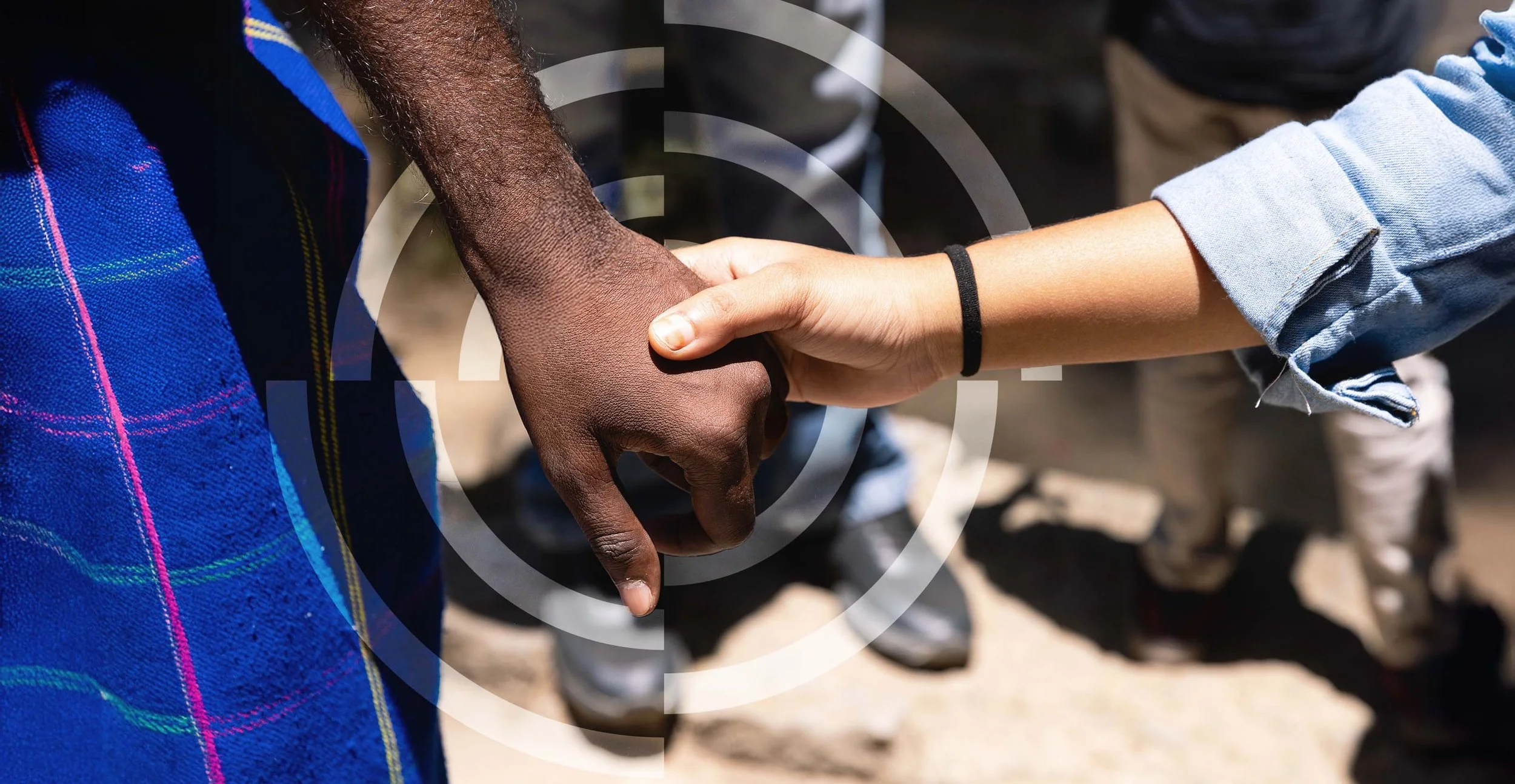 Two people holding hands, one with darker skin and wearing a blue plaid shirt, the other with lighter skin and wearing a denim shirt with a black hair tie on the wrist.