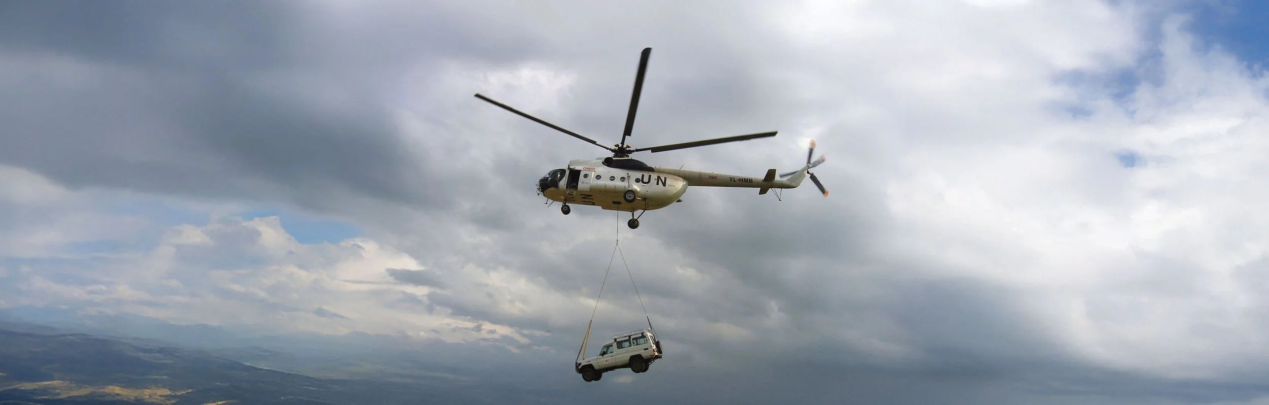 A helicopter flying in the sky hauling a vehicle beneath it with a cable, against a backdrop of cloudy sky and distant landscape.