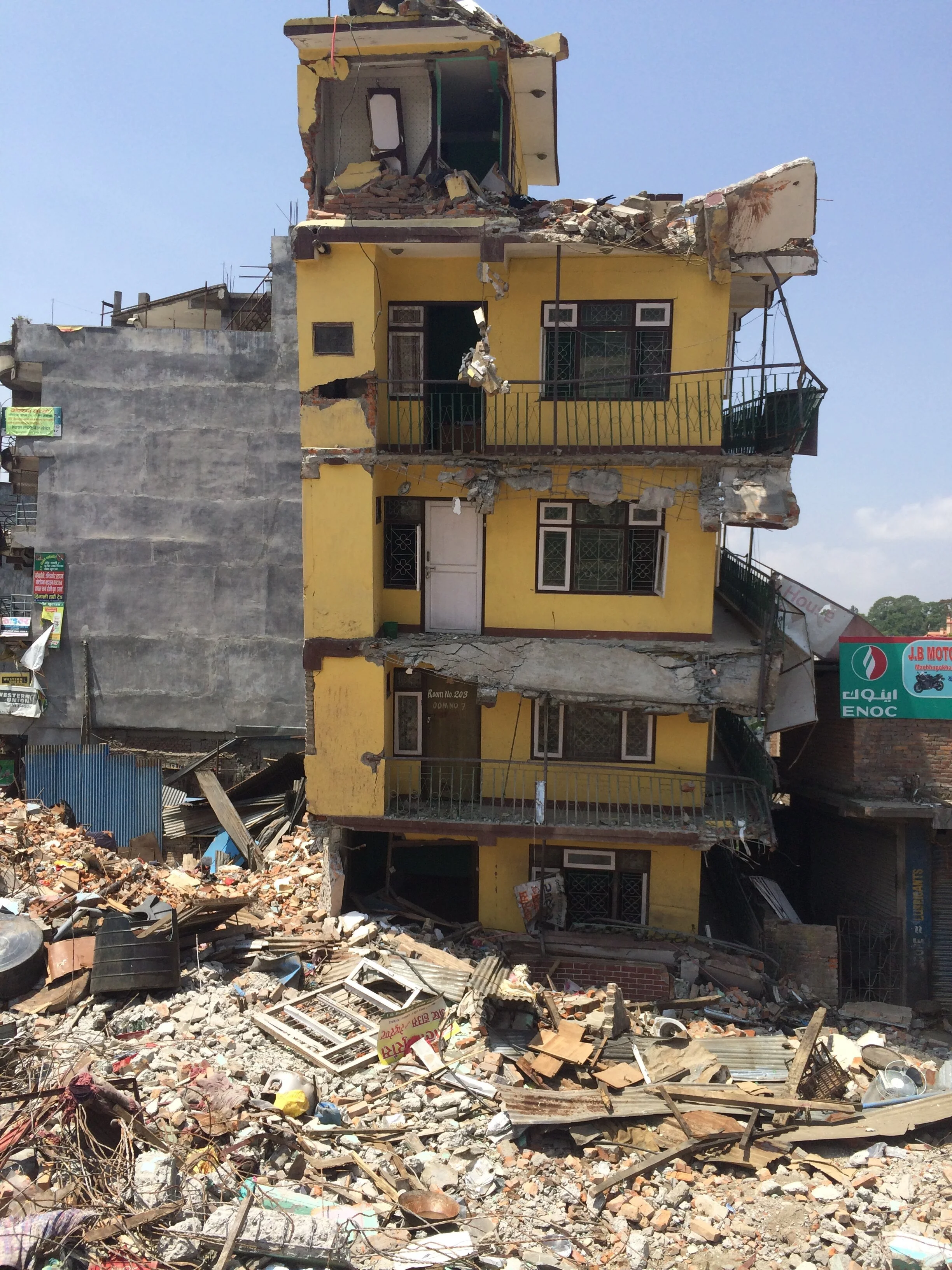 A multi-story yellow building with extensive damage from an earthquake, with debris and rubble scattered at its base and portions of the structure collapsed.