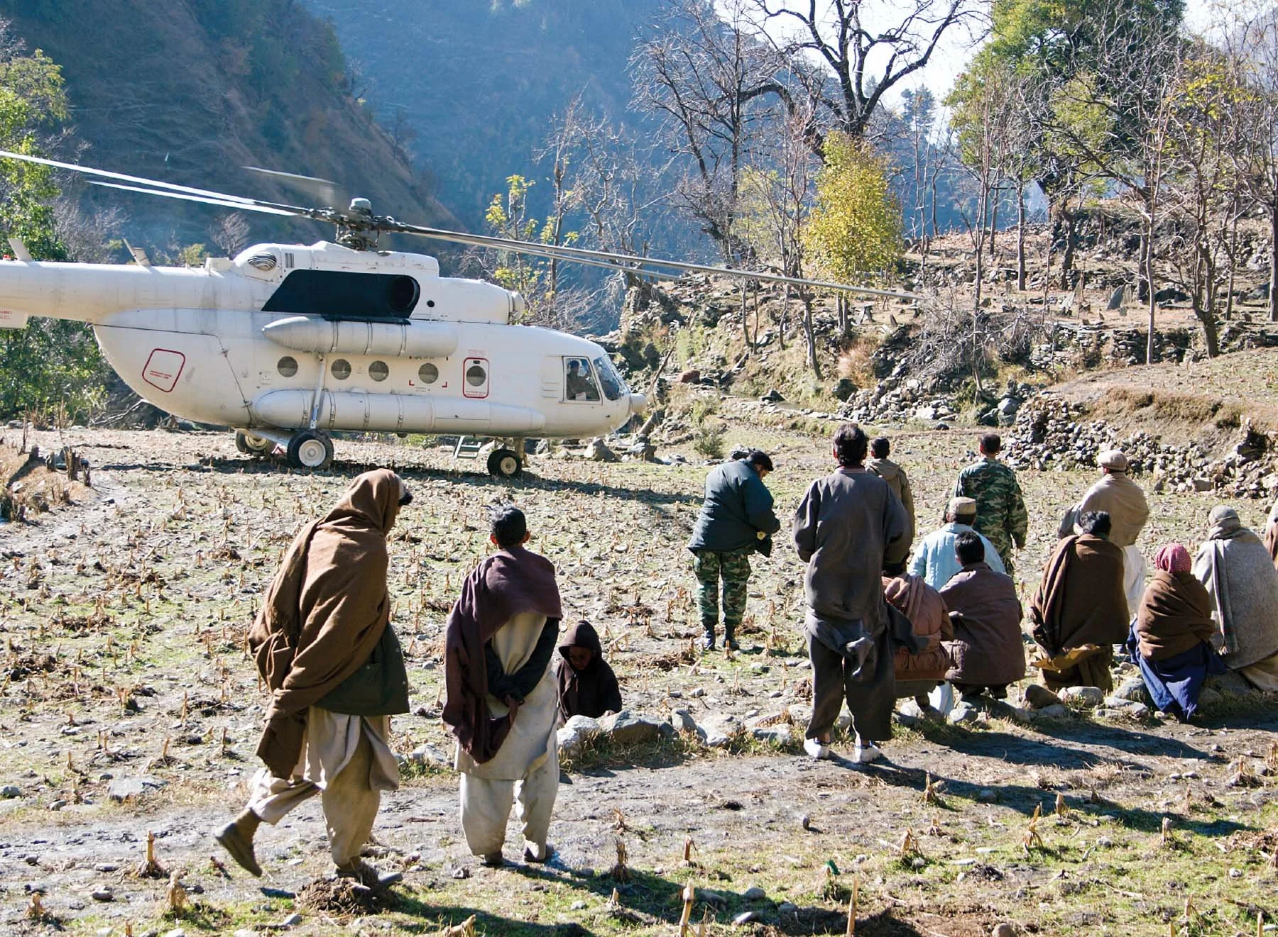 A rescue helicopter on the ground with a group of people, some sitting and some walking, in a mountainous, barren landscape with leafless trees.