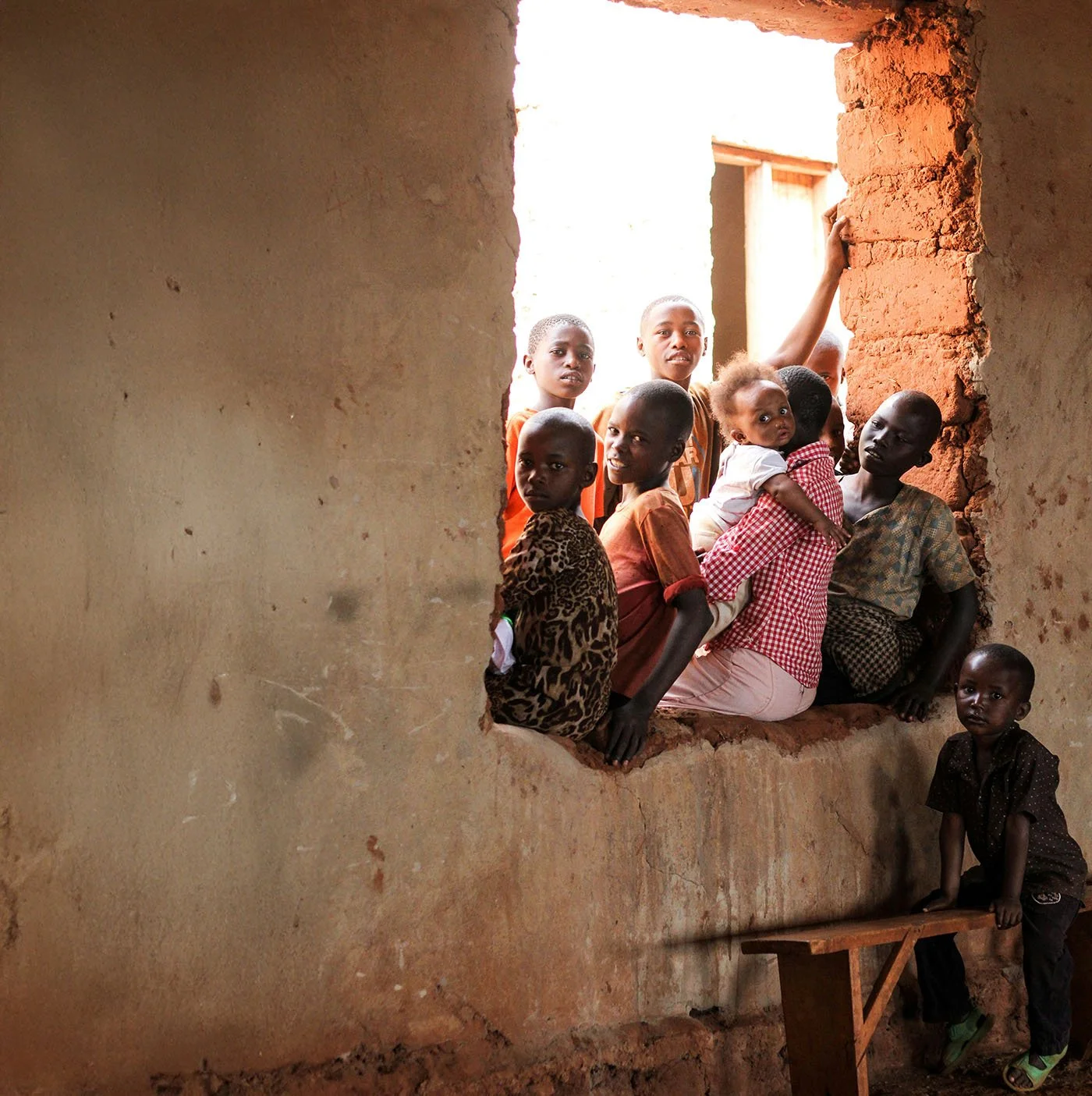 A group of children and young women sitting in a large, rectangular window opening in a mud-brick wall of a building with natural light coming in.
