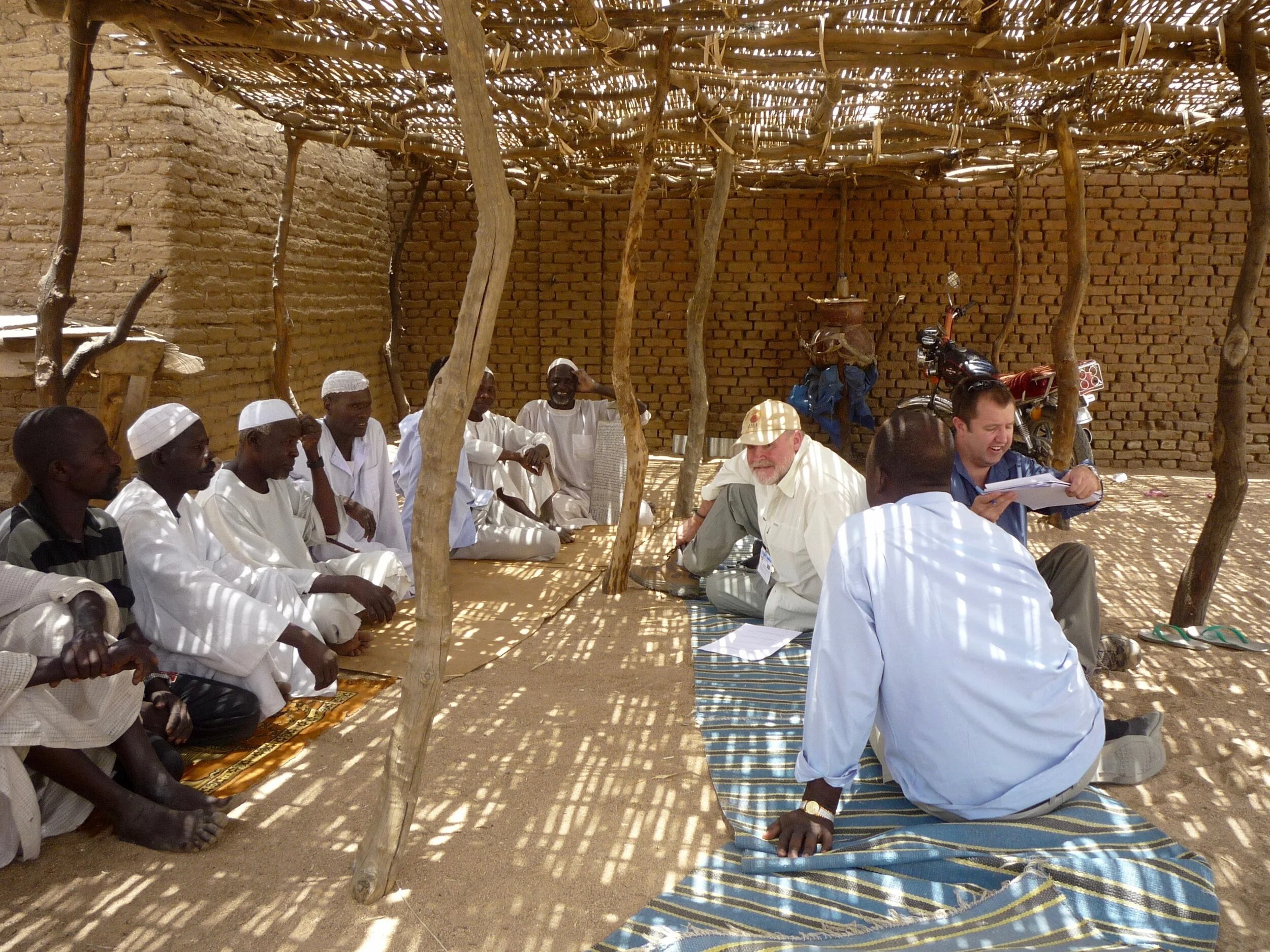 Group of men sitting on the ground inside a traditional shade structure, engaging in discussion, with some using notebooks and papers, in front of a brick wall, with motorcycles and bags in the background.