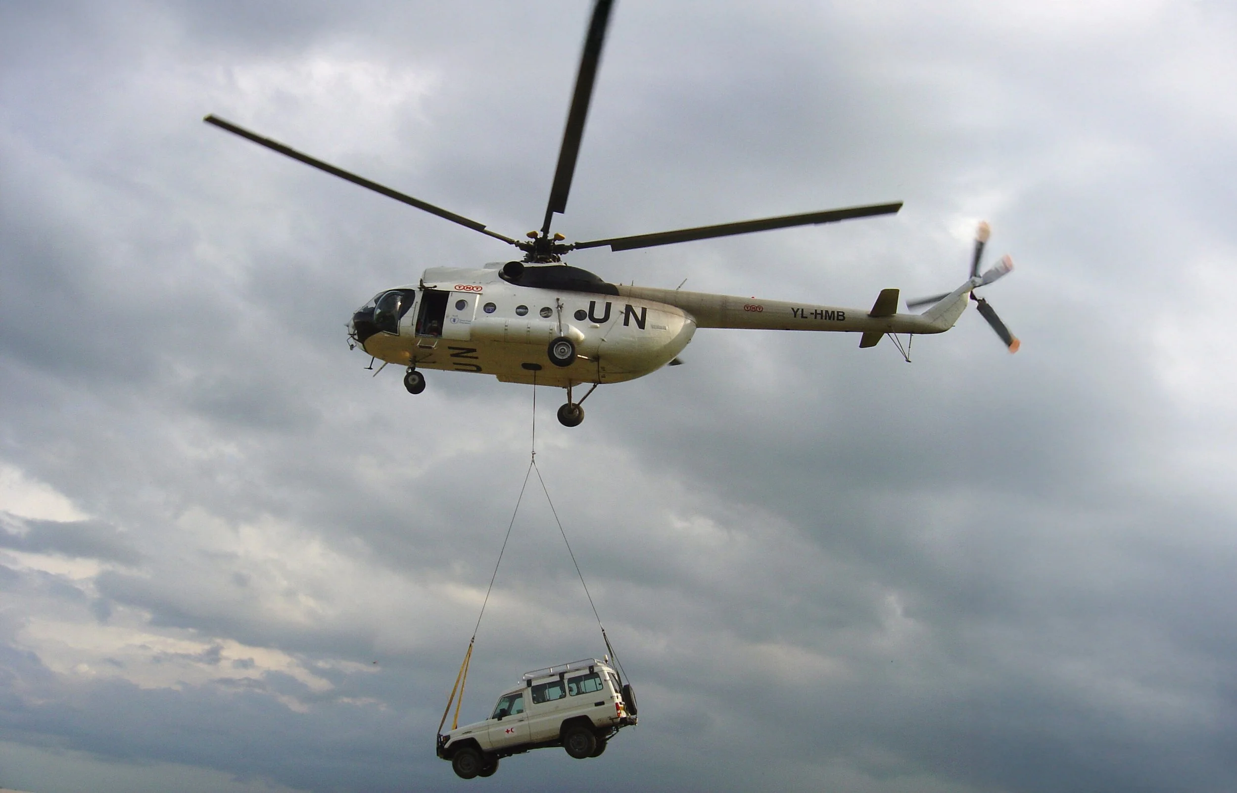 A helicopter is flying in the sky, lifting a white vehicle with red cross markings, attached by a cable, beneath it.
