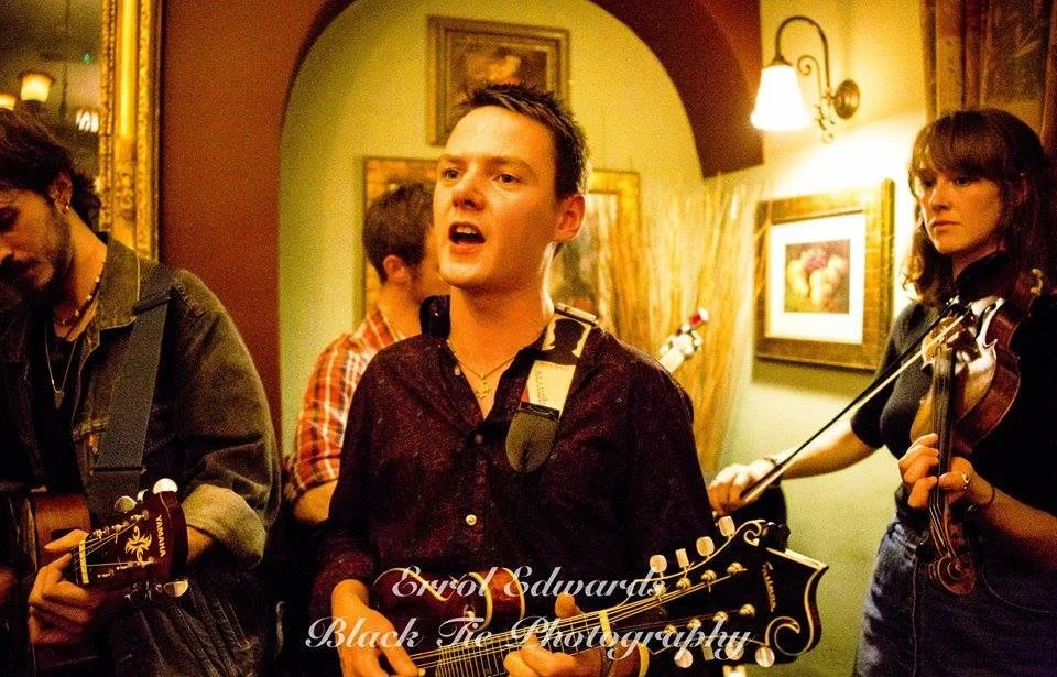 A group of musicians performing indoors, with a man in the center singing and playing guitar, surrounded by others with instruments, in a warmly lit room with paintings on the wall.