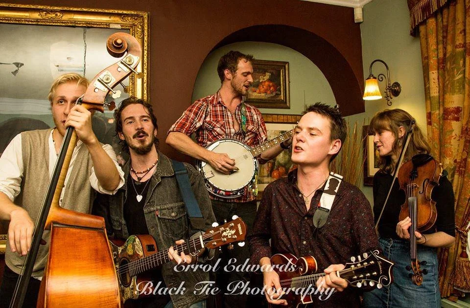 A group of five musicians playing instruments and singing together indoors. They include a standing bassist, a seated acoustic guitarist, a seated banjo player, a standing guitarist, and a standing violinist. The room has warm lighting, a mirror, and decorative curtains.