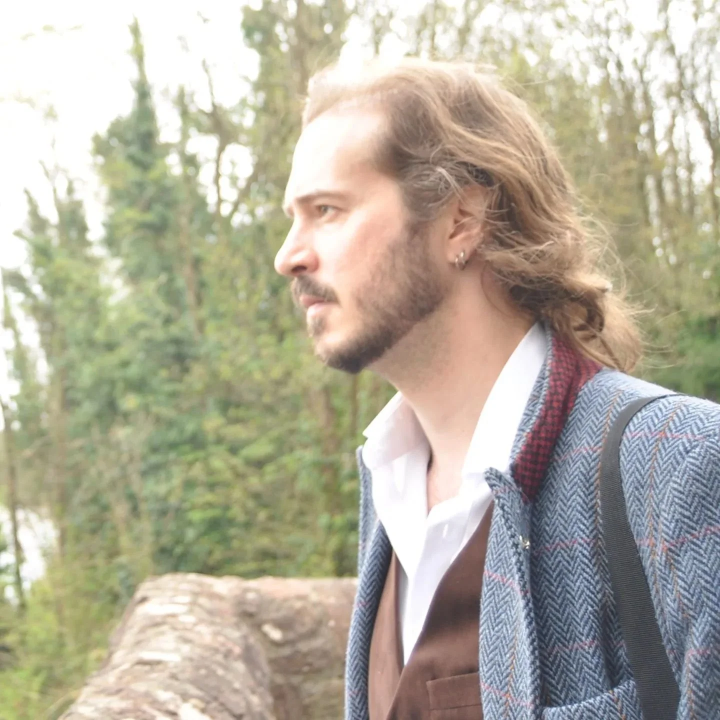 A man with long hair, beard, and earrings looking to the left outdoors with trees in the background.