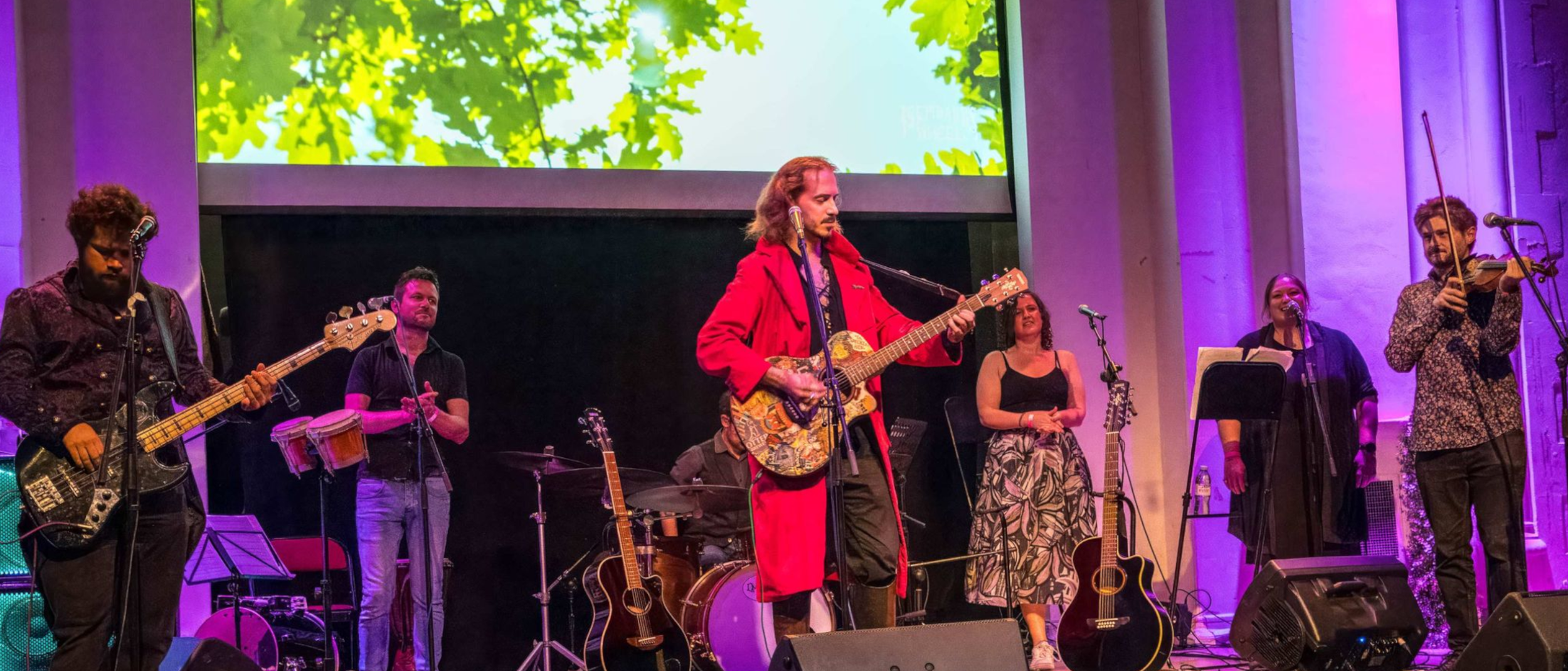 Musicians performing on stage with various instruments including guitars, violin, drums, and percussion, with a large screen showing green leafy trees in the background.