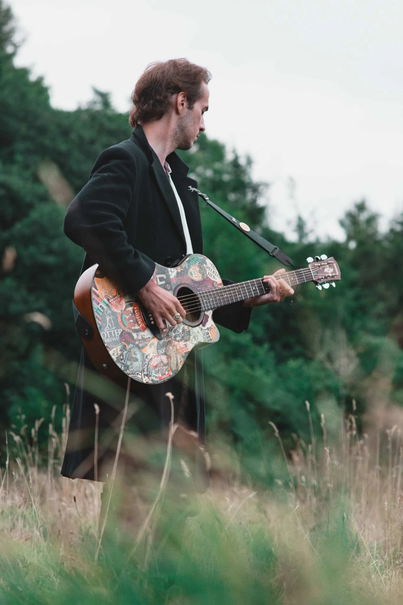 A man playing an acoustic guitar with a sticker-covered body outdoors in a field with tall grass, trees, and an overcast sky.