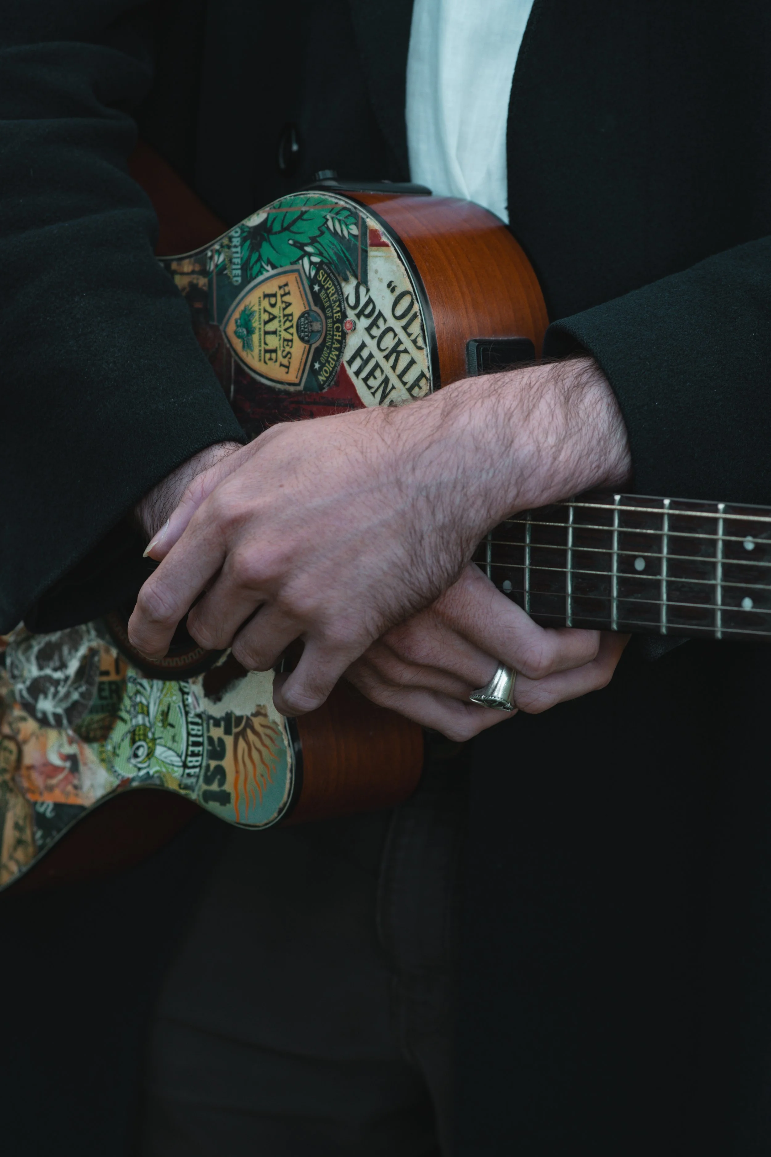 Close-up of a person holding an acoustic guitar with a decorative pickguard, with their hands overlapping, wearing a wedding ring, in a black jacket and white shirt.