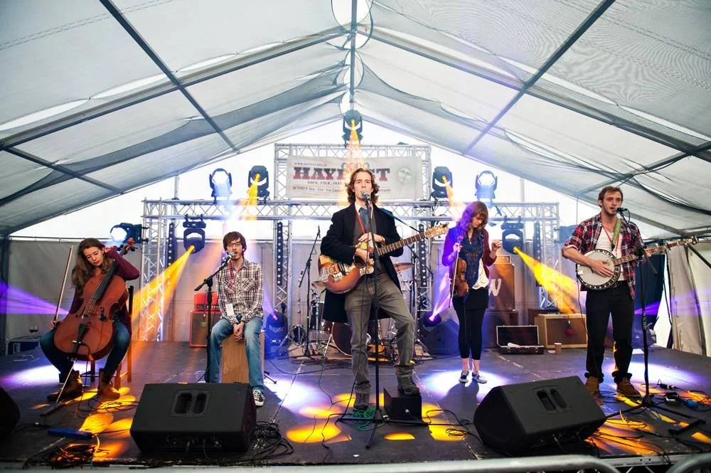 A band performing on stage inside a large tent at a music festival called Hayloft, with five members playing instruments and singing, surrounded by stage lights.