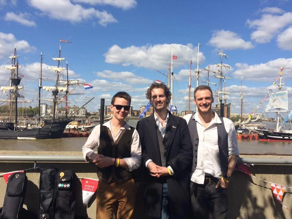 Three men stand in front of a harbor with tall ships under a partly cloudy sky.