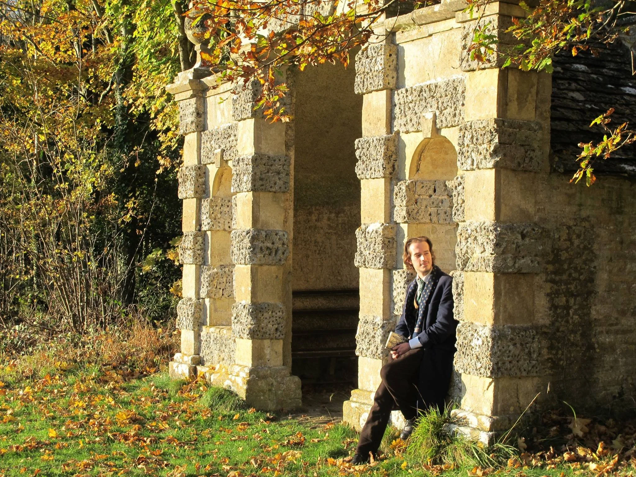 Man sitting on a stone ledge beside an old stone building with an archway, surrounded by autumn leaves and foliage.
