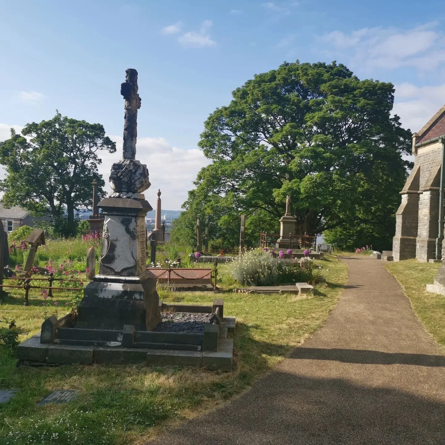 Graveyard with a stone cross monument, trees, flowers, and a dirt pathway on a sunny day with a blue sky.