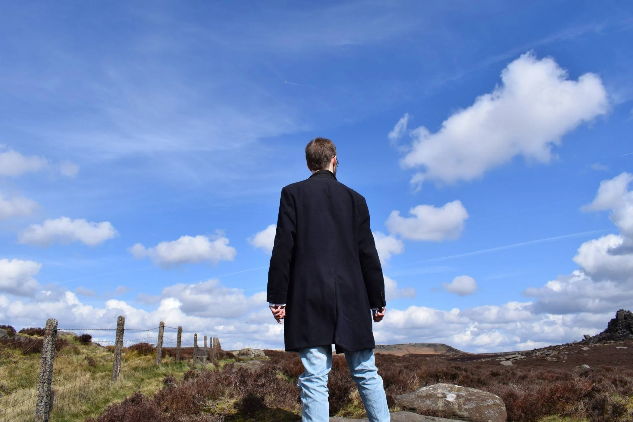 A man dressed in a long dark coat and light blue jeans standing outdoors on rocky terrain with purple and green vegetation, looking towards a blue sky with scattered white clouds.